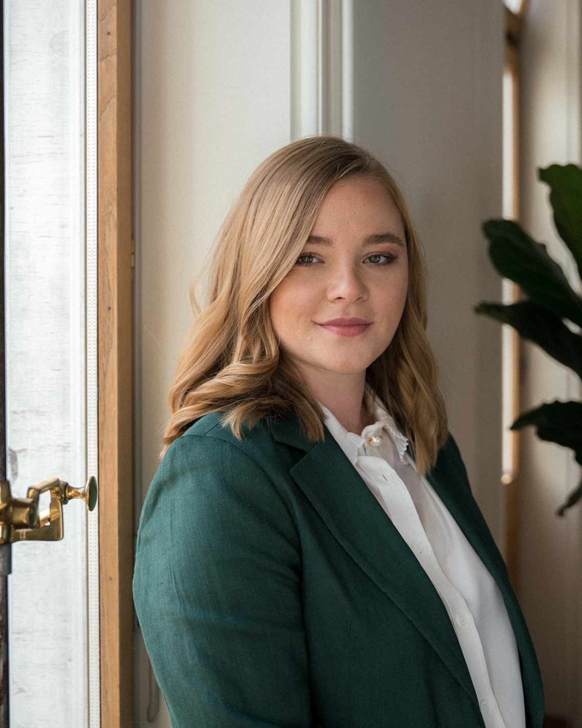Jeune femme avec des cheveux blonds bouclés, portant un tailleur vert foncé et une chemise blanche, debout près d'une fenêtre intérieure, regardant vers l'objectif.