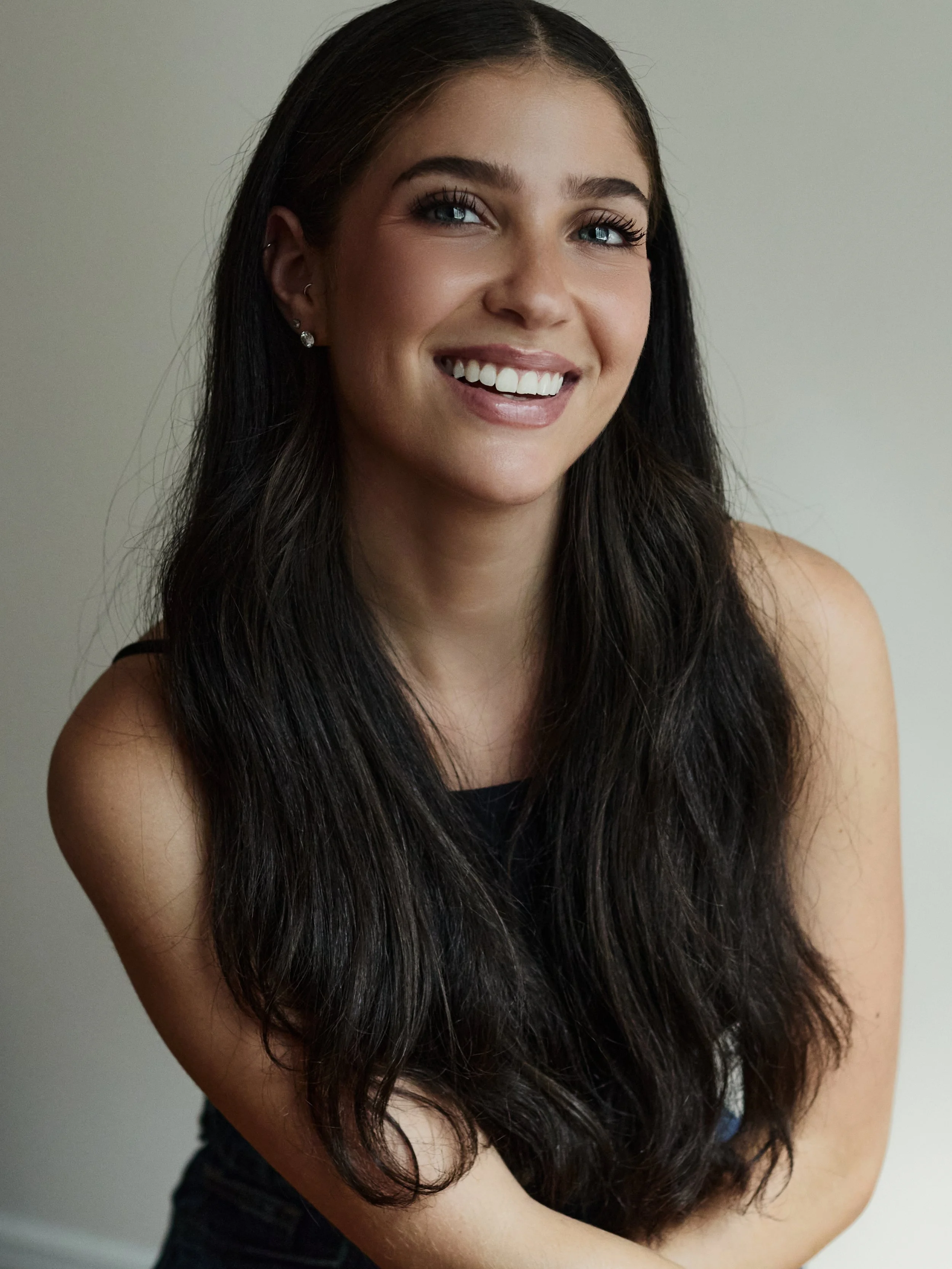 Smiling woman with long dark hair, wearing earrings, posing with crossed arms against a neutral background.
