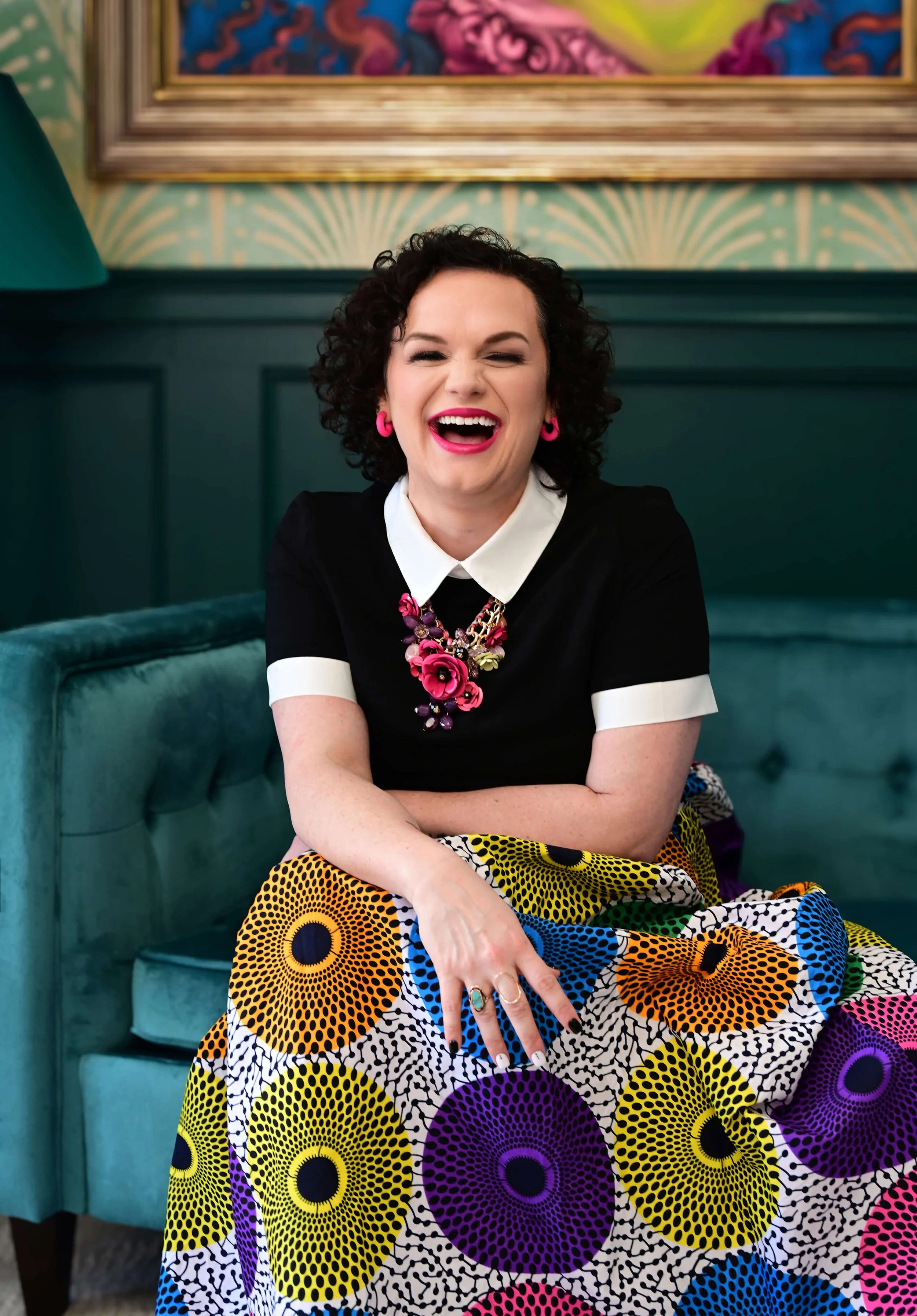 A woman with curly dark hair, wearing bright pink earrings and a black top with a white collar, smiling and laughing with eyes closed. She sits on a teal cushioned bench with her arms crossed, wearing a colorful, patterned skirt with large circles in yellow, purple, and black. In the background, there is a framed colorful painting on the wall.