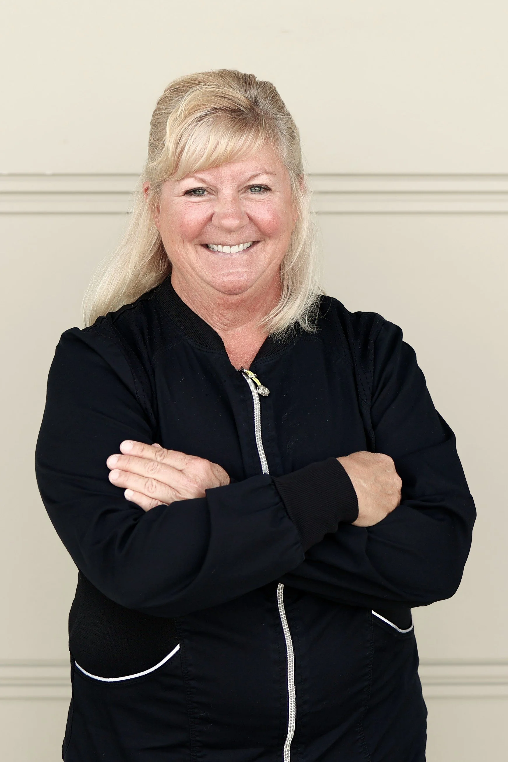 A woman with blonde hair wearing a black zip-up jacket standing against a plain beige wall, smiling with her hands in her pockets.