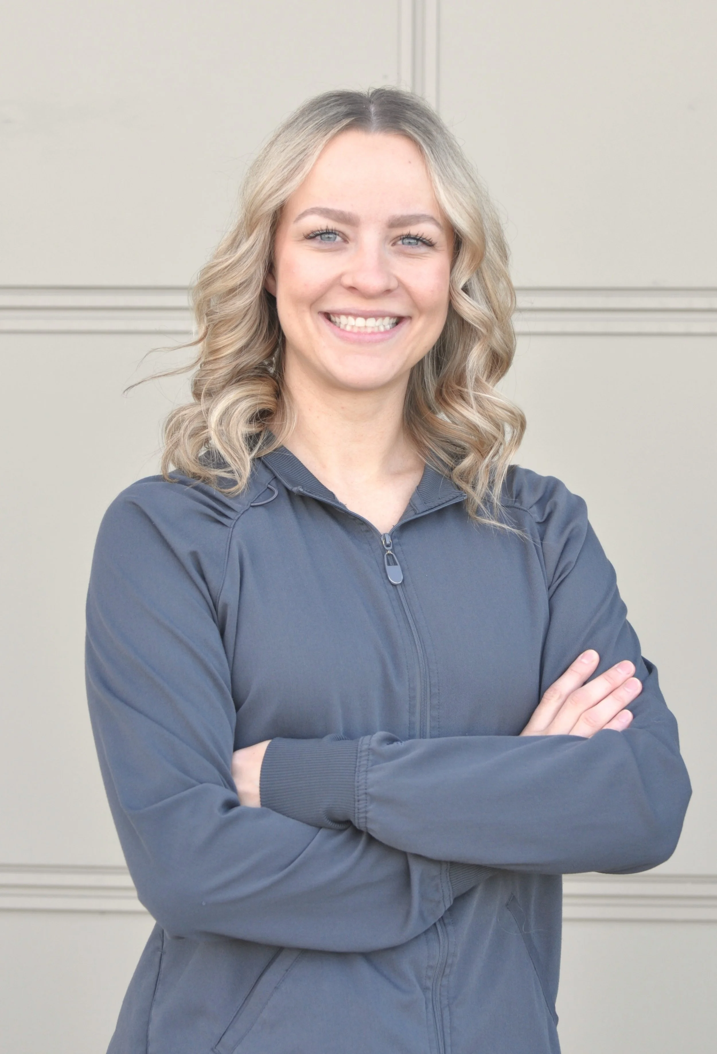 A woman with blonde hair smiling and crossing her arms, wearing a gray zip-up jacket, standing against a plain beige wall.