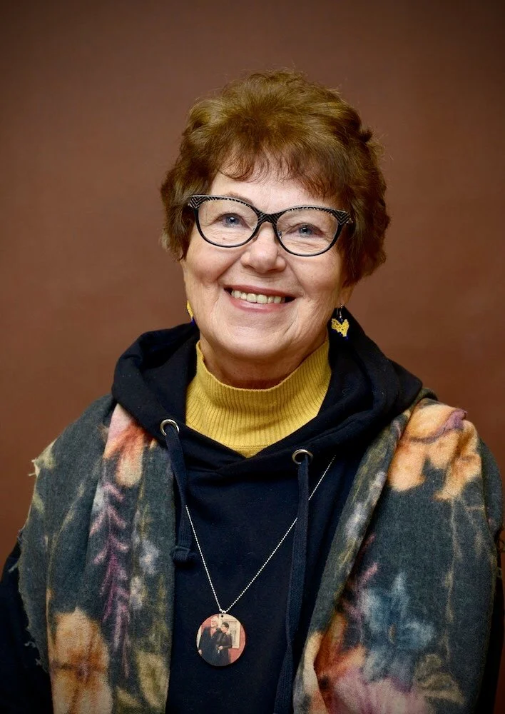 Vanhempi henkilö hymyilee, yllään mustat silmälasit, keltainen paita ja värikäs huivi. Tausta on ruskea.