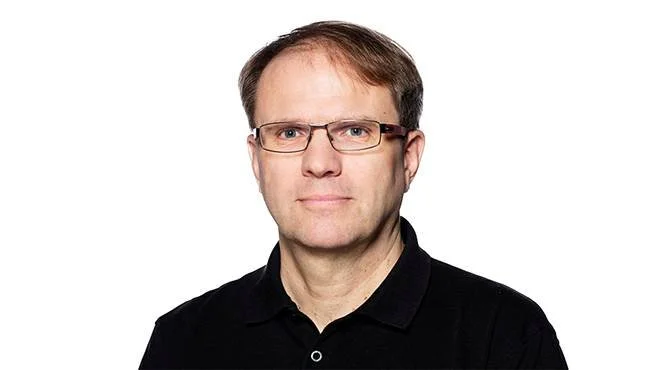 Mies, jolla on silmälasit, tumma paita, katsoo suoraan kameraan.