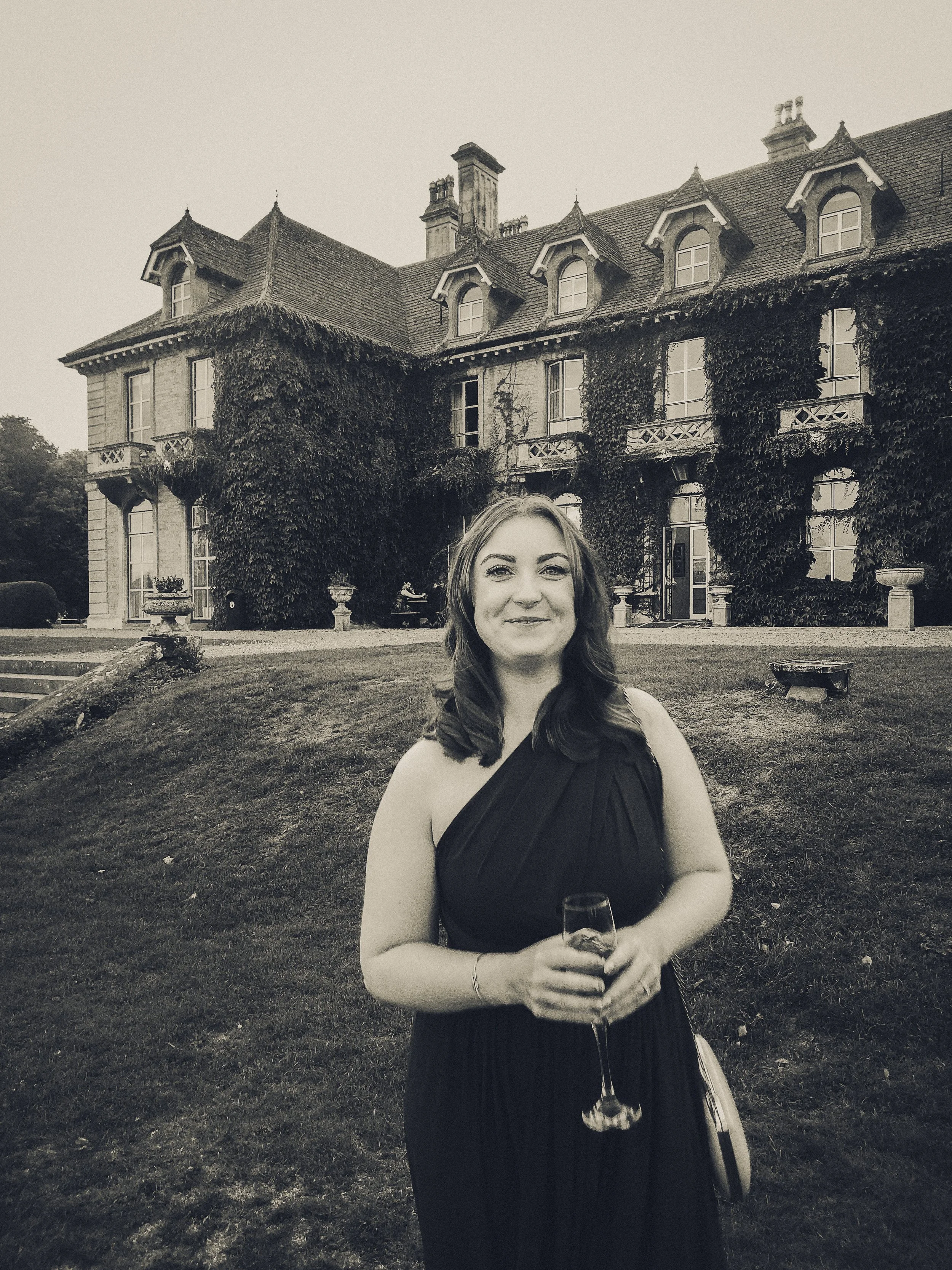 A woman in a black dress holding a glass of wine, standing outside a large, ivy-covered mansion.
