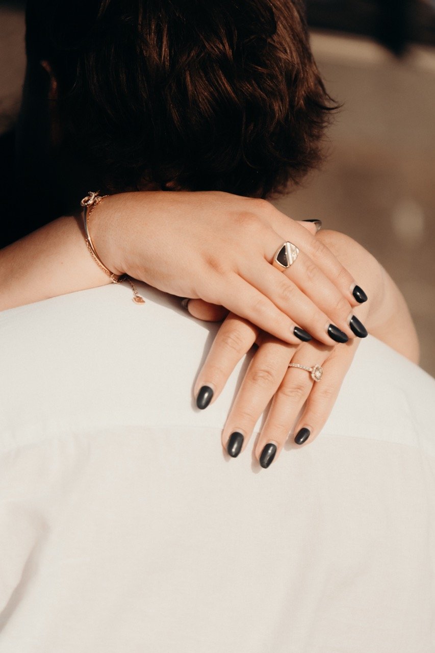 Close-up of a woman's hands resting on a white surface, showing black nail polish, rings, and a bracelet.