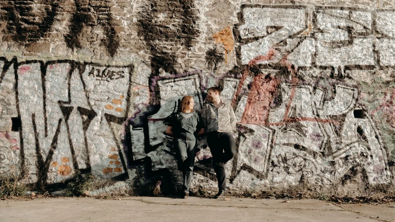 A man and woman standing and leaning against a graffiti-covered brick wall, looking at each other, in an urban setting.