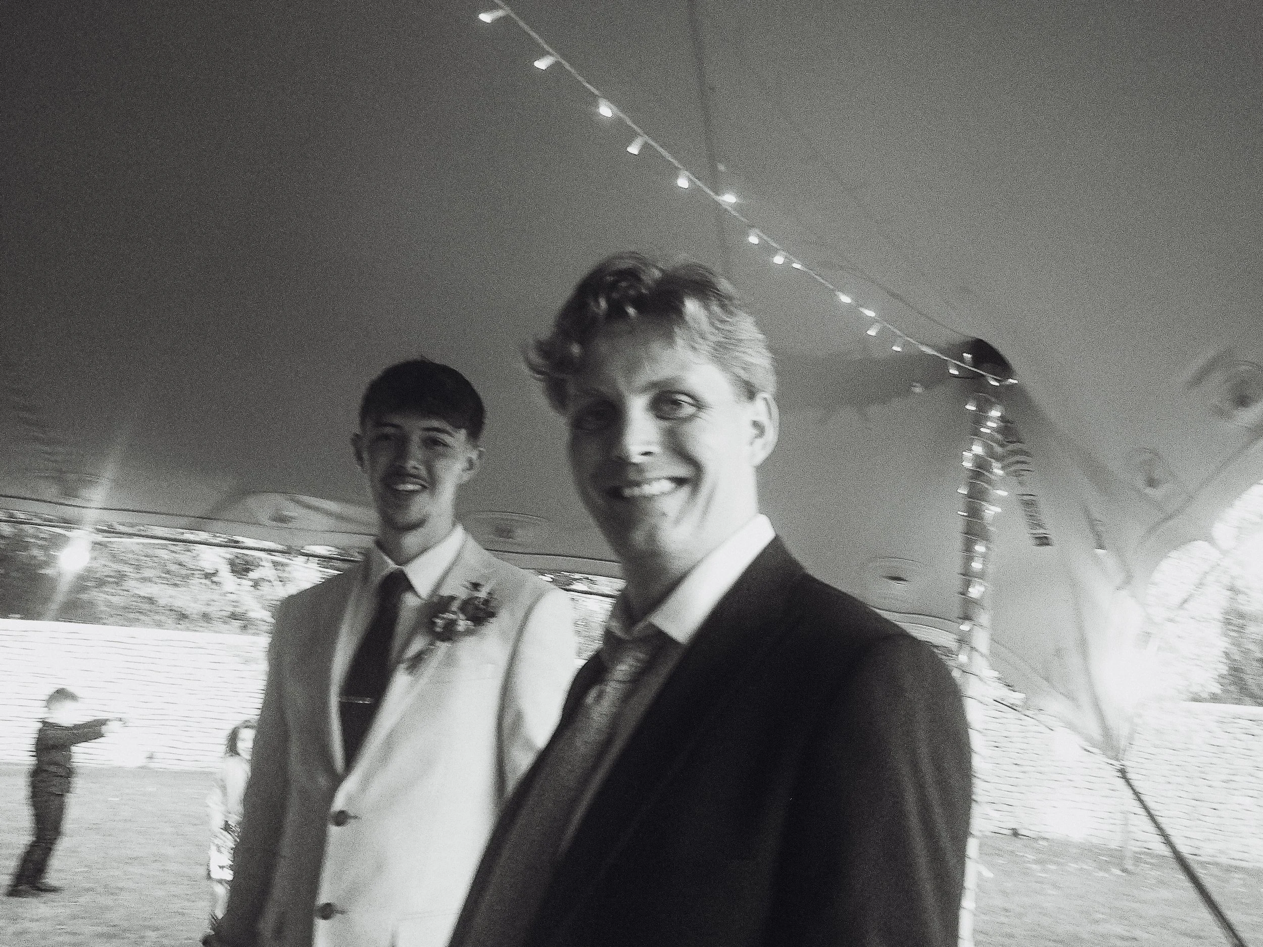 Two young men in tuxedos smiling, standing inside a decorated tent, at an outdoor event.