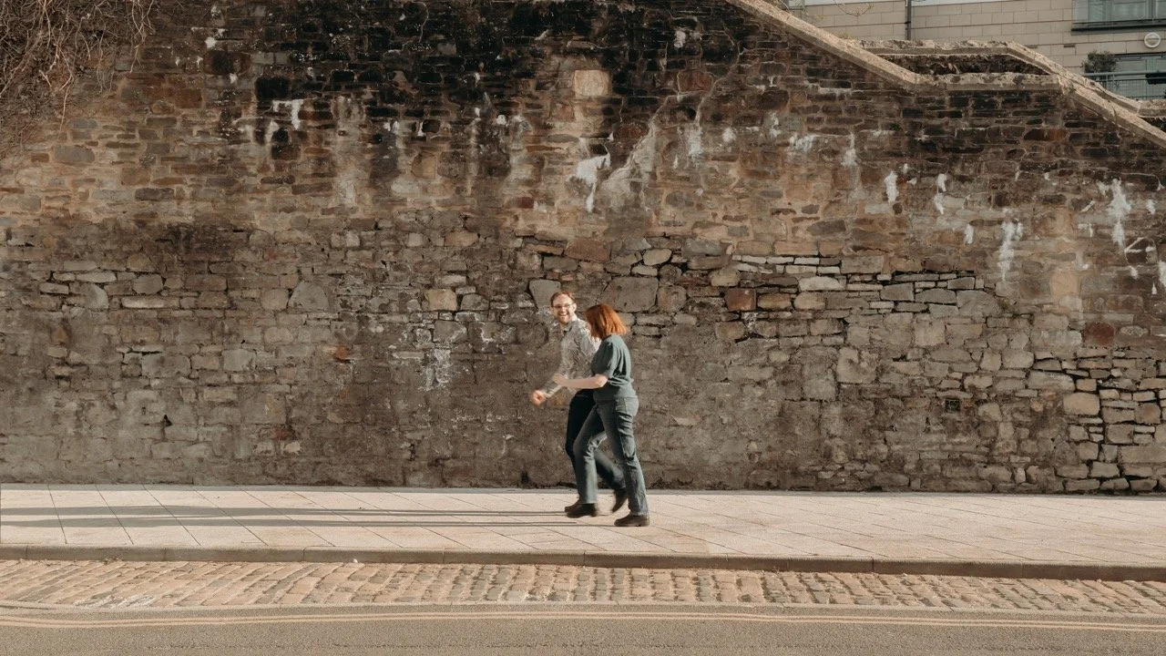 Two people walking along a sidewalk next to an old brick wall with some white paint streaks.