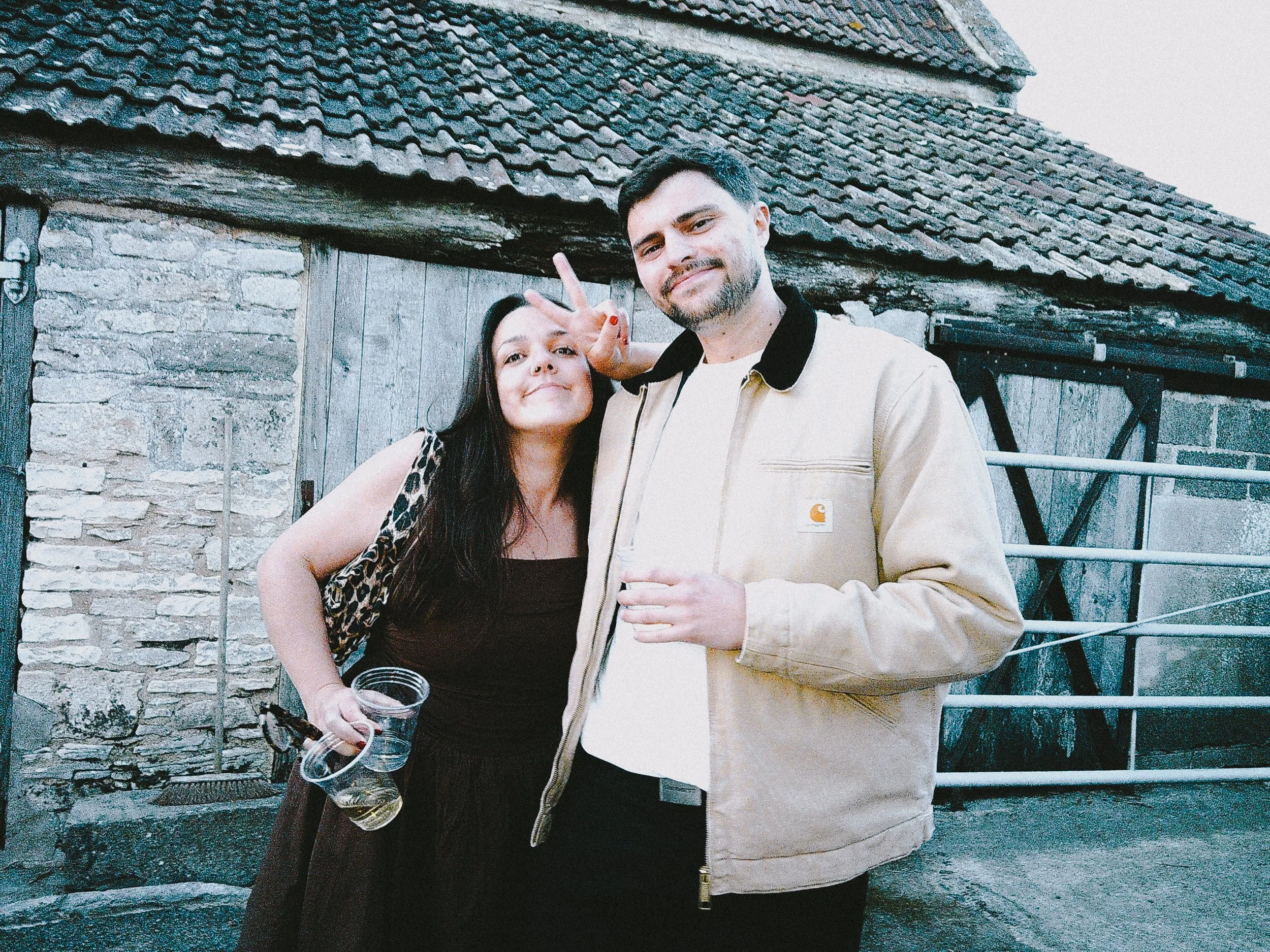 A young woman with long dark hair, wearing a dark dress and leopard print cardigan, holding a plastic cup, making a peace sign next to a young man with short dark hair and beard, wearing a beige Carhartt jacket, both smiling outdoors in front of a ru