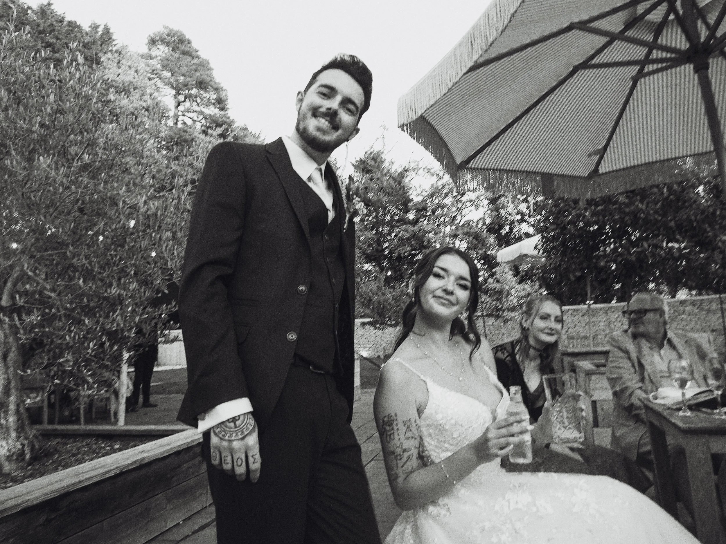 A black and white photo of four people at an outdoor gathering. One man in a suit and tie is standing and smiling, while three women sit at a table with drinks. The woman in the middle is wearing a wedding dress and has tattoos on her arms. An umbrel