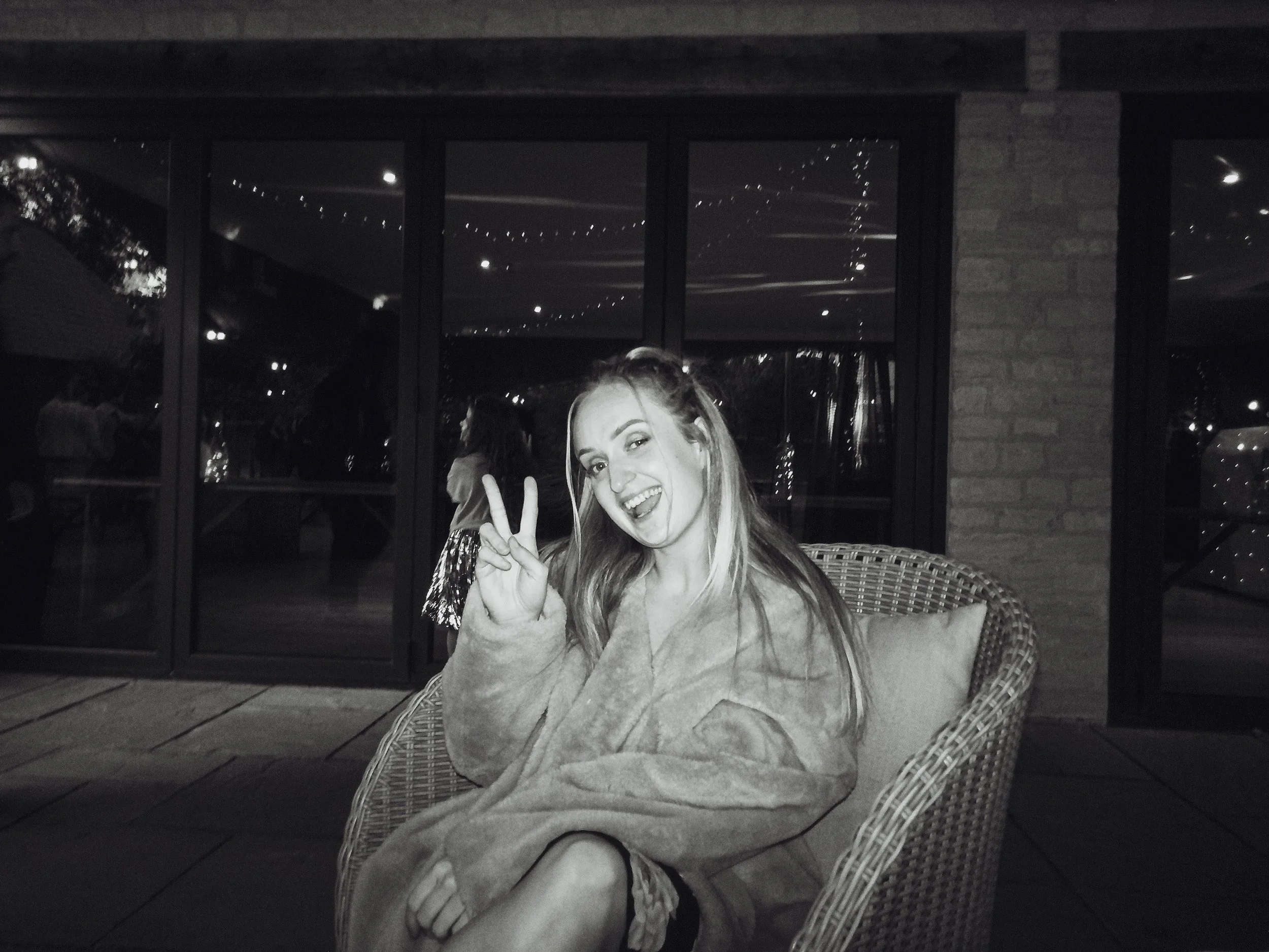 A young woman sitting in a wicker chair outdoors at night, making a peace sign and tongue-out pose, smiling and wearing a bathrobe, with reflections of string lights visible in the glass behind her.