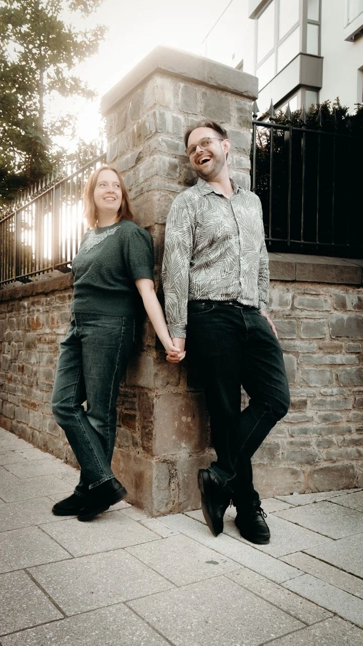 A man and woman holding hands and leaning against a brick corner wall outdoors, smiling at each other with a tree and building in the background, sunlight behind them.