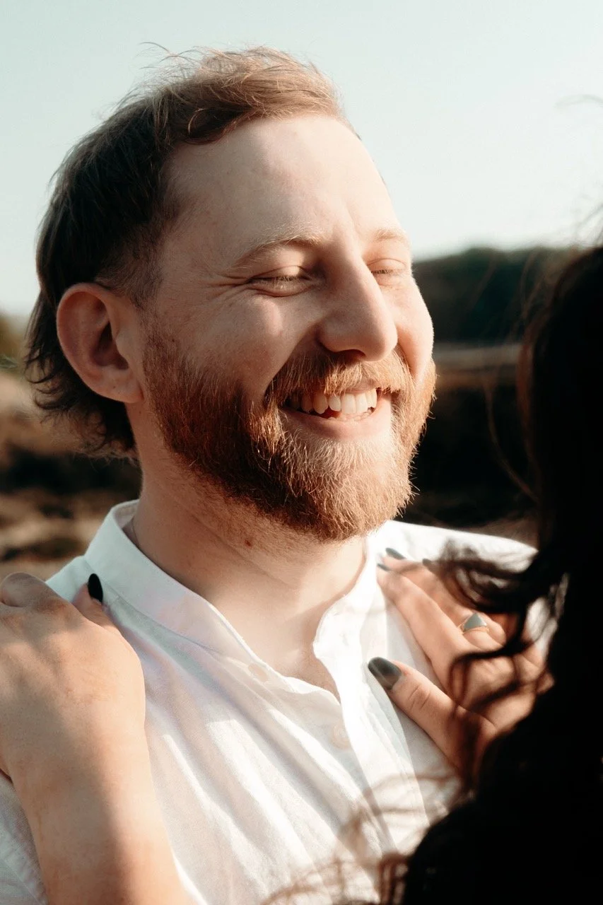 A man with a beard and closed eyes smiling, being embraced by a woman with dark hair and manicured nails.