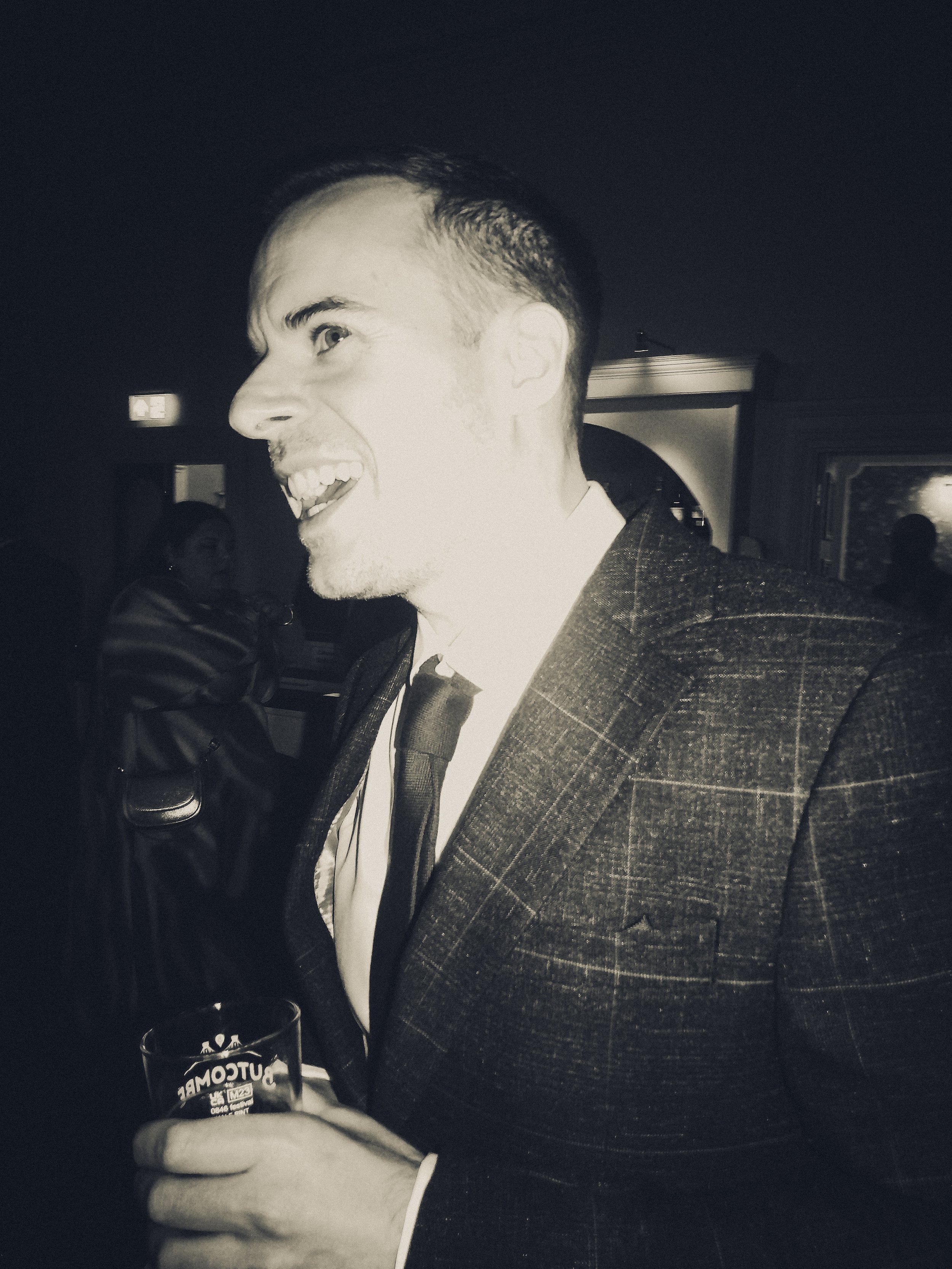 A young man with dark hair, styled short on the sides, smiling and holding a glass of beer, wearing a patterned suit and tie at a social gathering indoors.