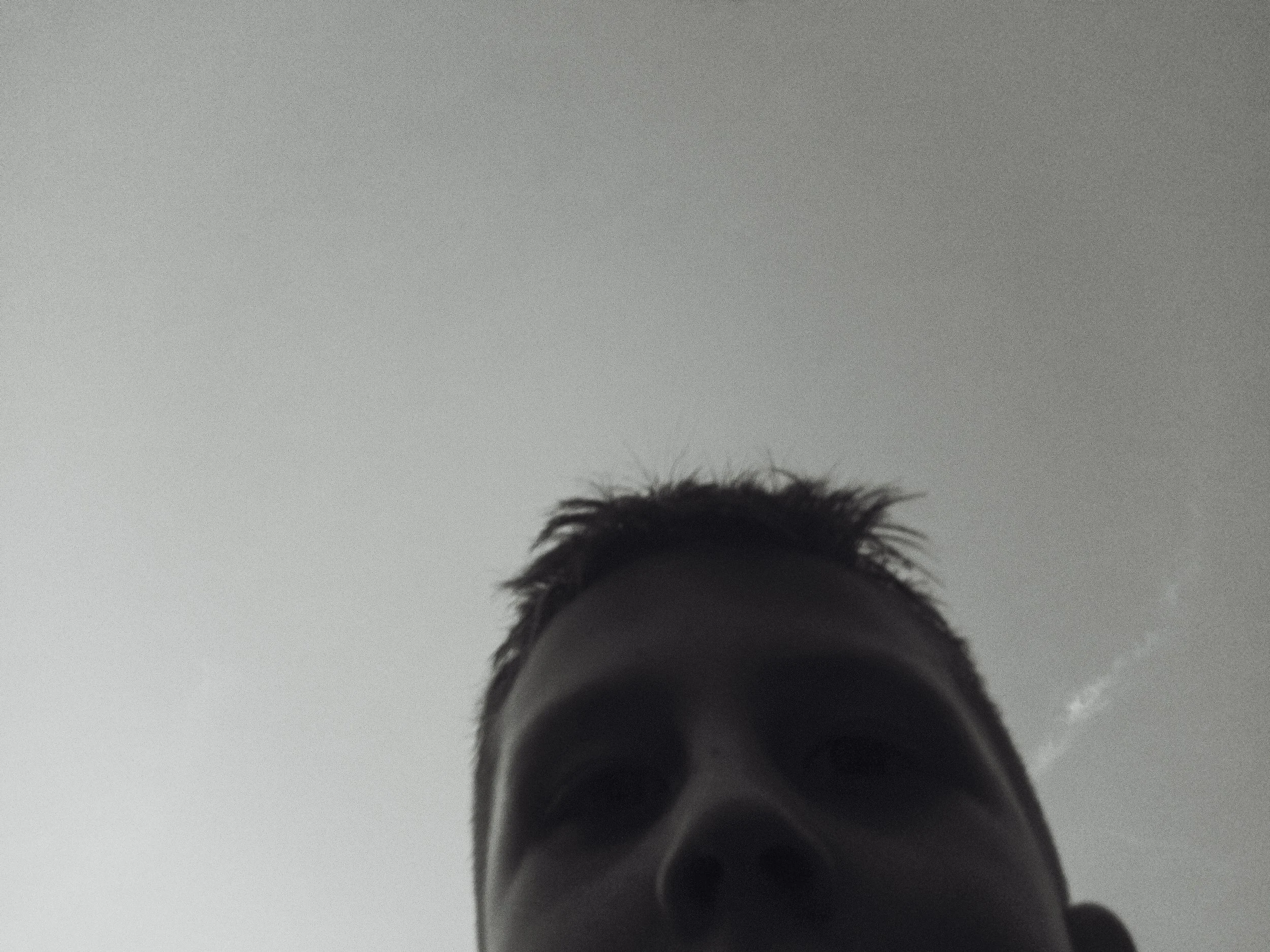 A black and white photo of a person's face taken from below, showing their nose, mouth, and eyes, with short hair, against a cloudy sky.