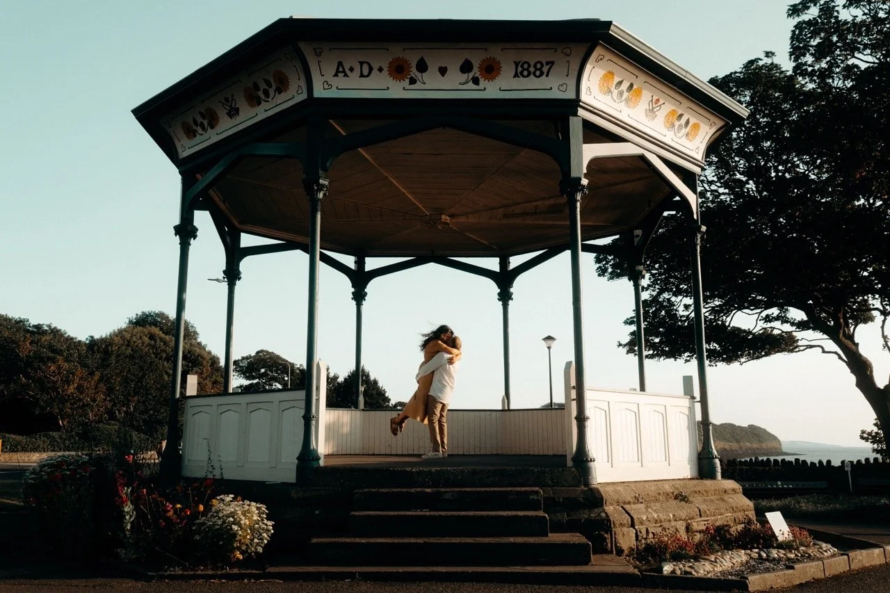 A couple embracing and hugging on a gazebo by the waterfront during sunset, with trees and a distant coastline visible.