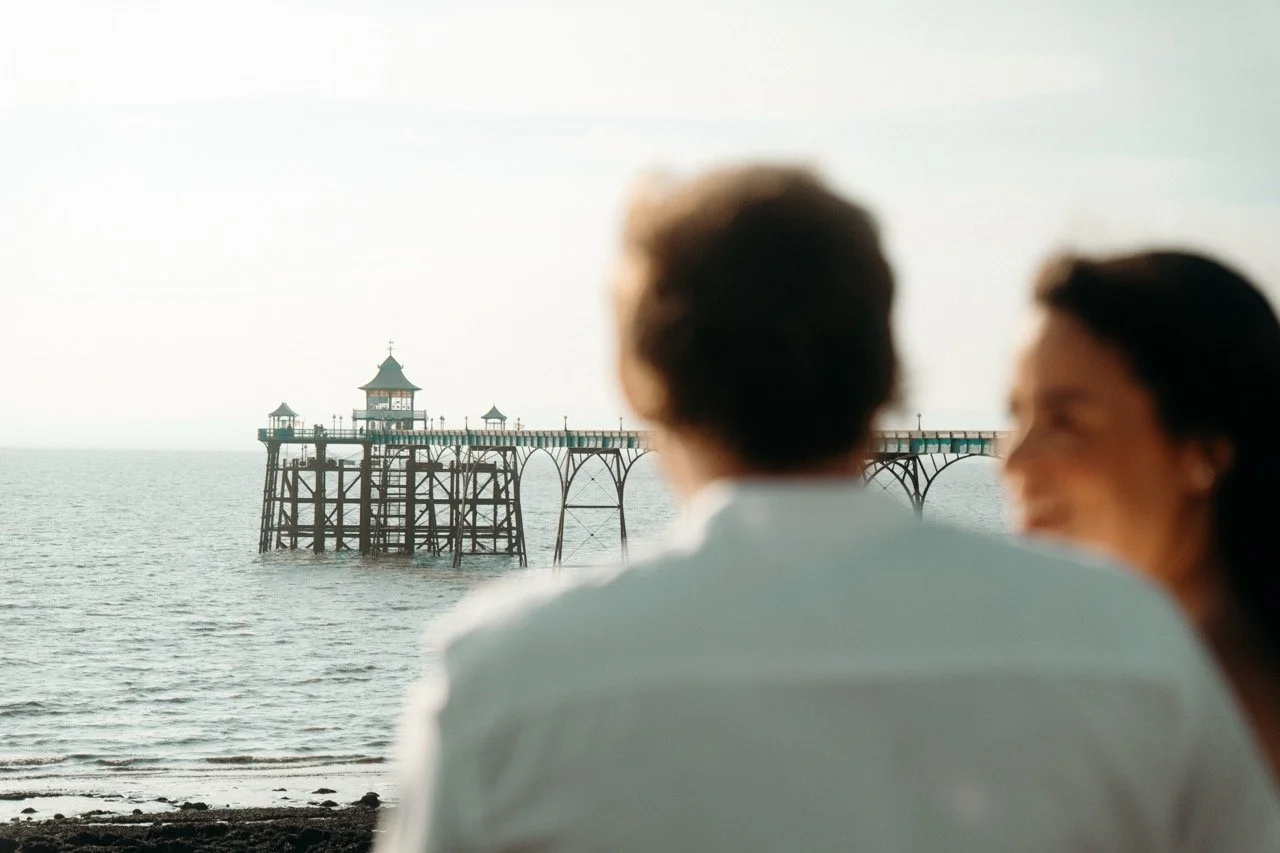 Blurred image of a man and woman sitting on the beach, with a pier extending into the ocean in the background.