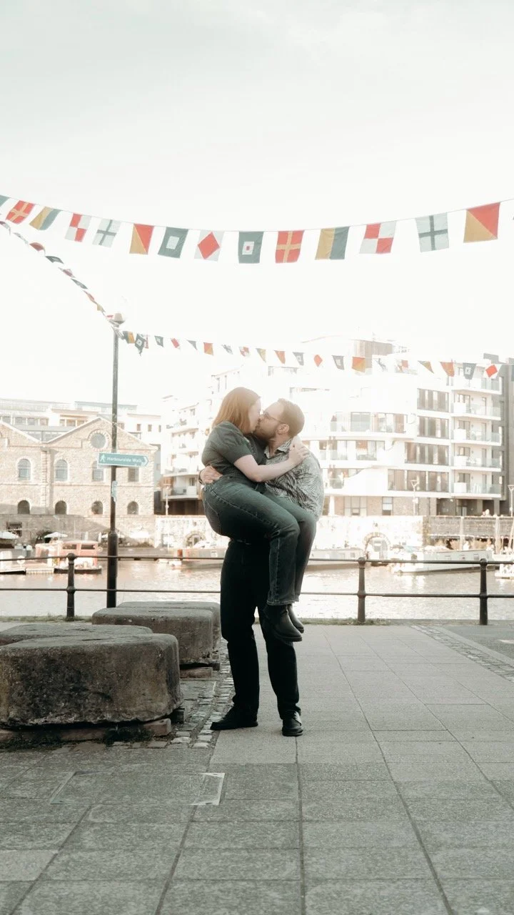 A couple sharing a kiss outdoors near a waterfront, with flags hanging above them and modern buildings in the background.