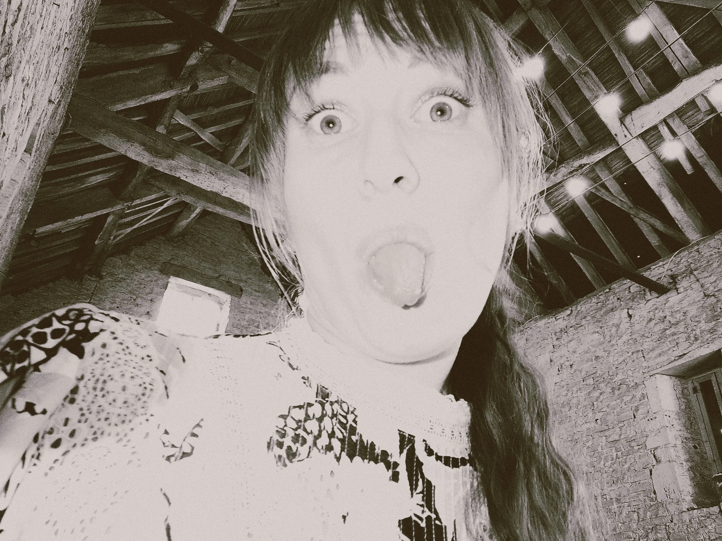 A woman with wide eyes and an open mouth making an expression of surprise or shock inside a rustic wooden building with exposed beams and a brick wall.