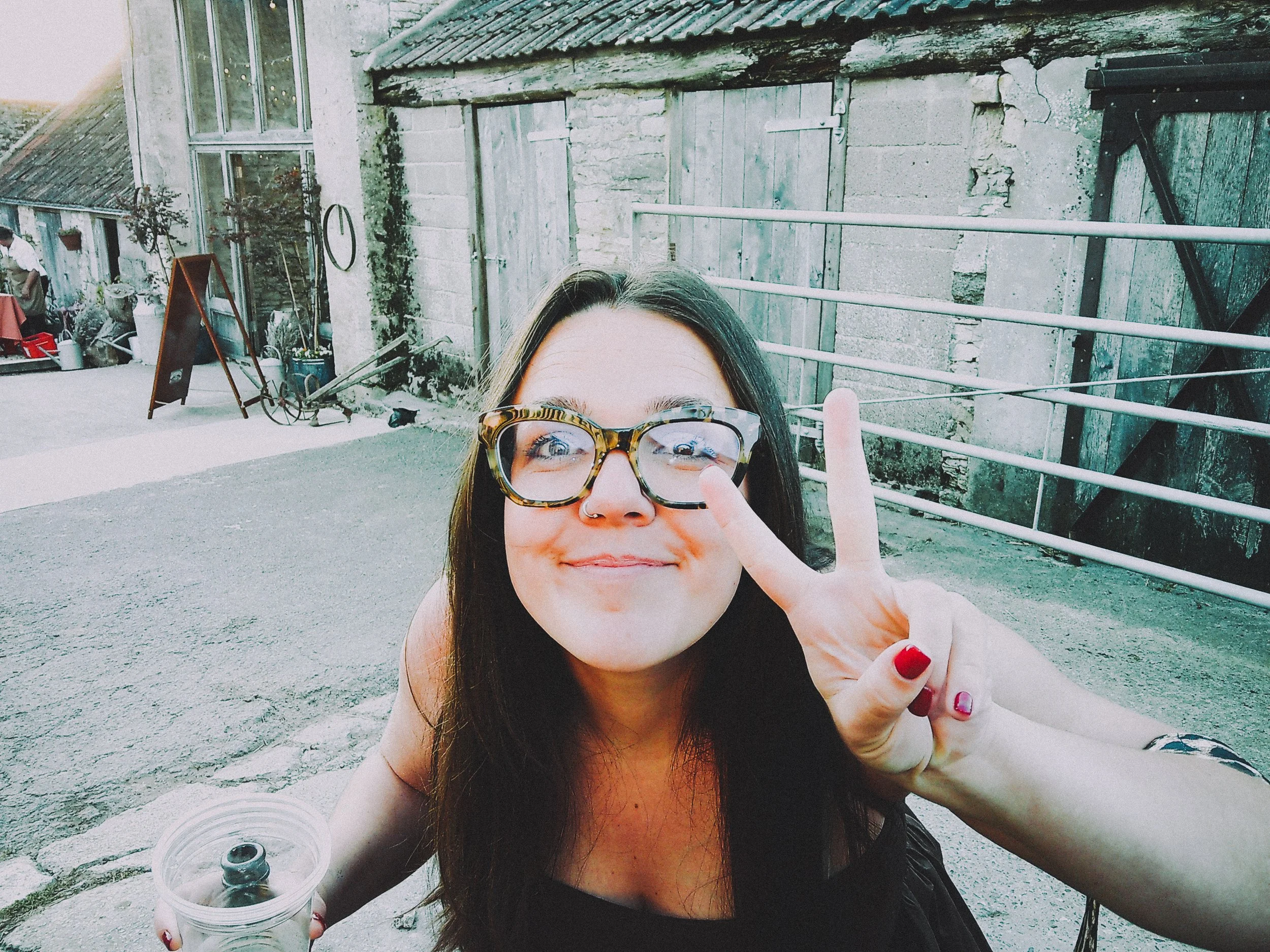 A woman with long dark hair, glasses, and red nails making a peace sign while smiling outdoors near an old stone and wood building.
