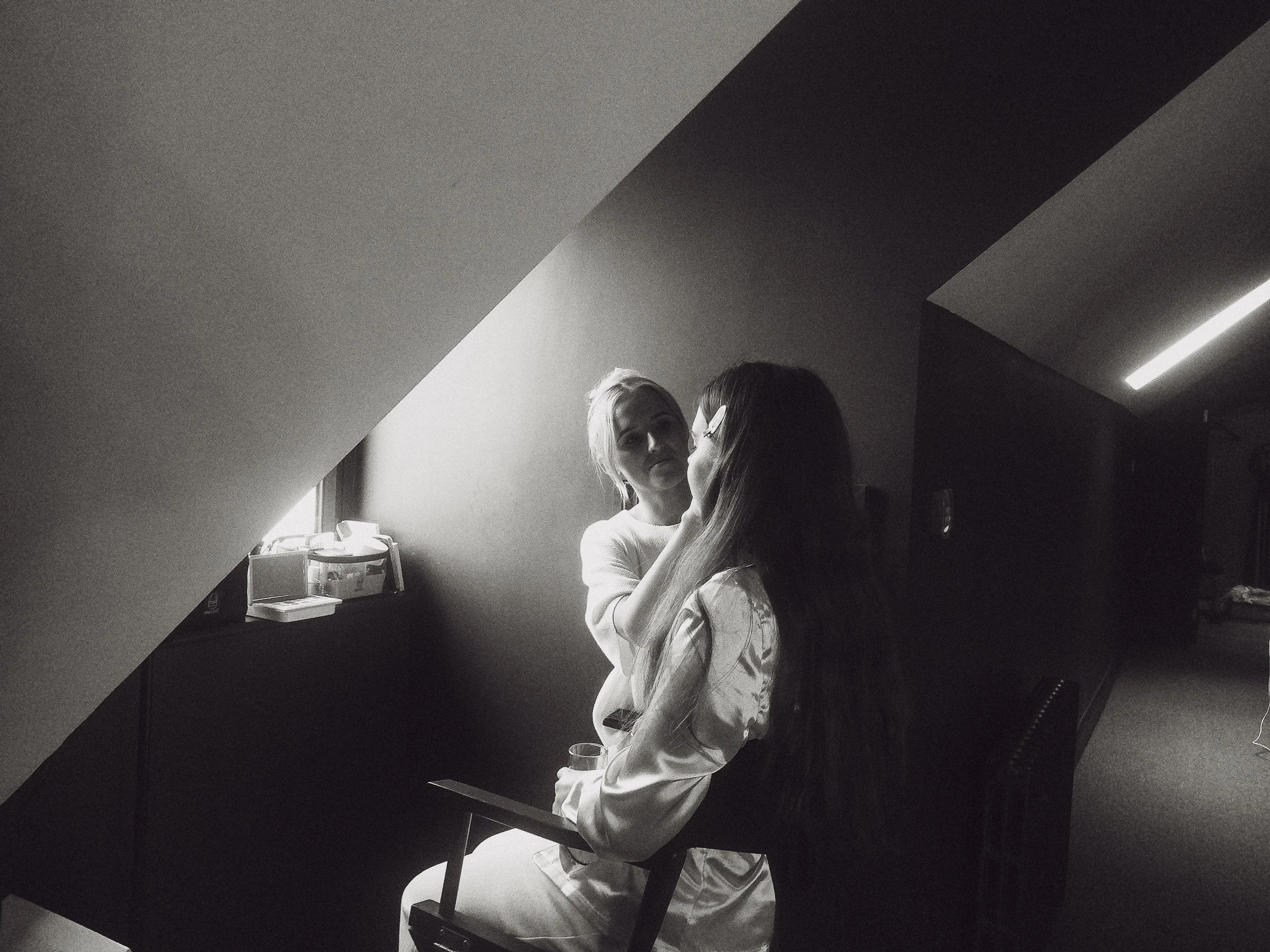 Two women, one with light hair and the other with dark hair and glasses, engaging in a close moment in a room with a sloped ceiling and a small window, illuminated by natural light.