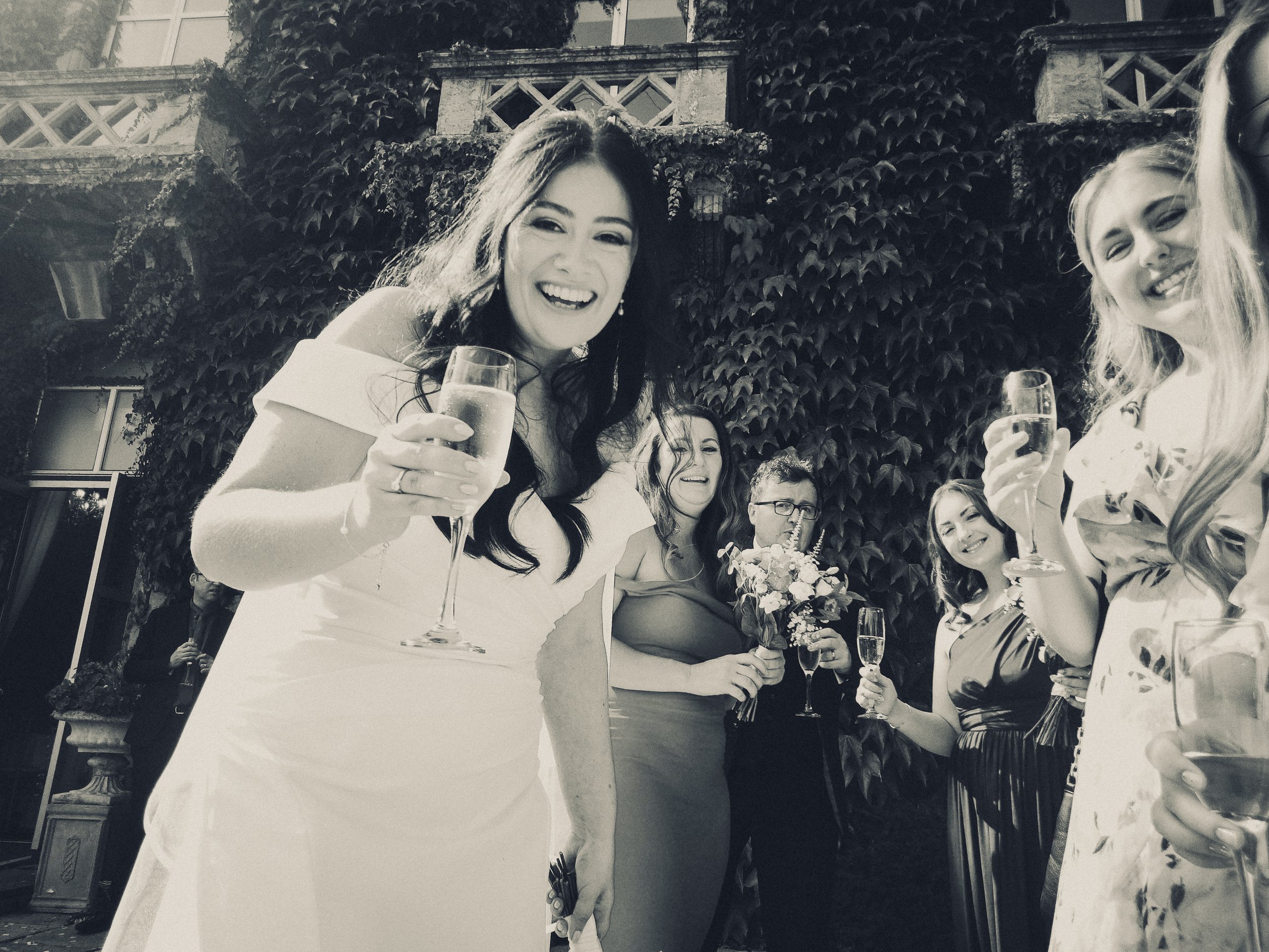 Group of women at a celebration holding glasses of champagne, smiling outdoors with a vine-covered building in the background.