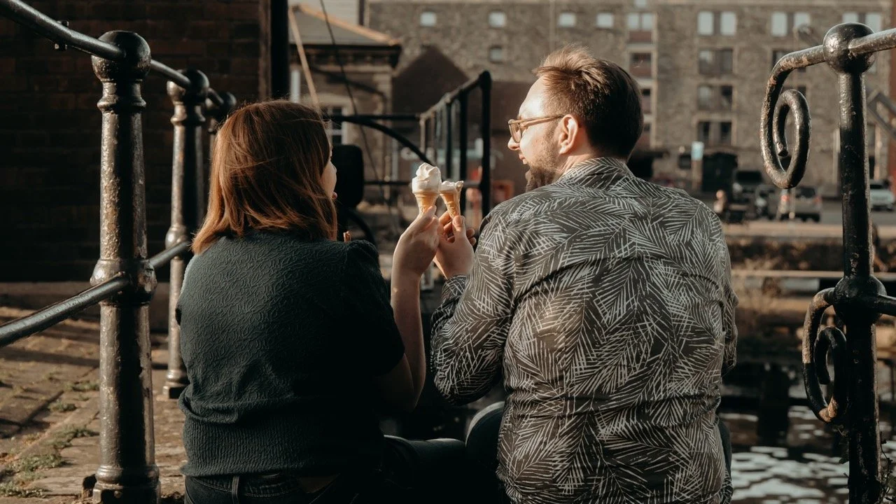 A man and woman sitting on a dock near water, sharing ice cream cones in the late afternoon with buildings in the background.