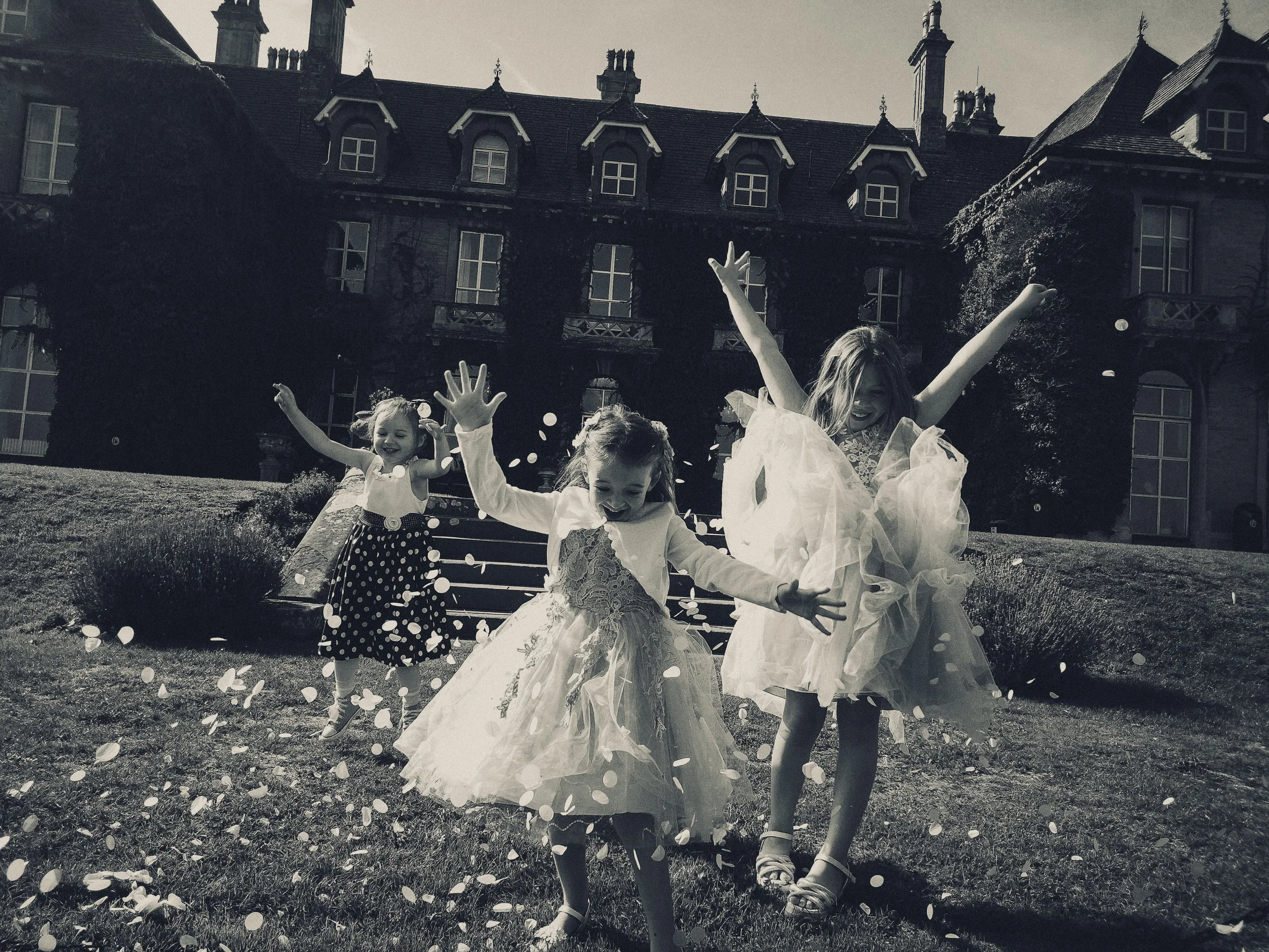 Three young girls in party dresses playing and throwing confetti outside in front of a large building with multiple windows and a slate roof.