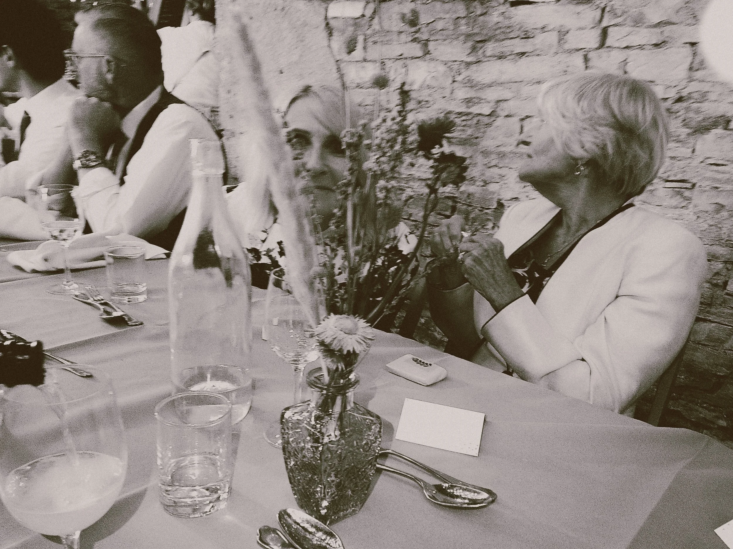 A woman with short, light-colored hair wearing a white blazer, sitting at a table with a flower arrangement, drinking from a glass, and looking to her right at an older woman with short, wavy hair wearing earrings and a white blazer.
