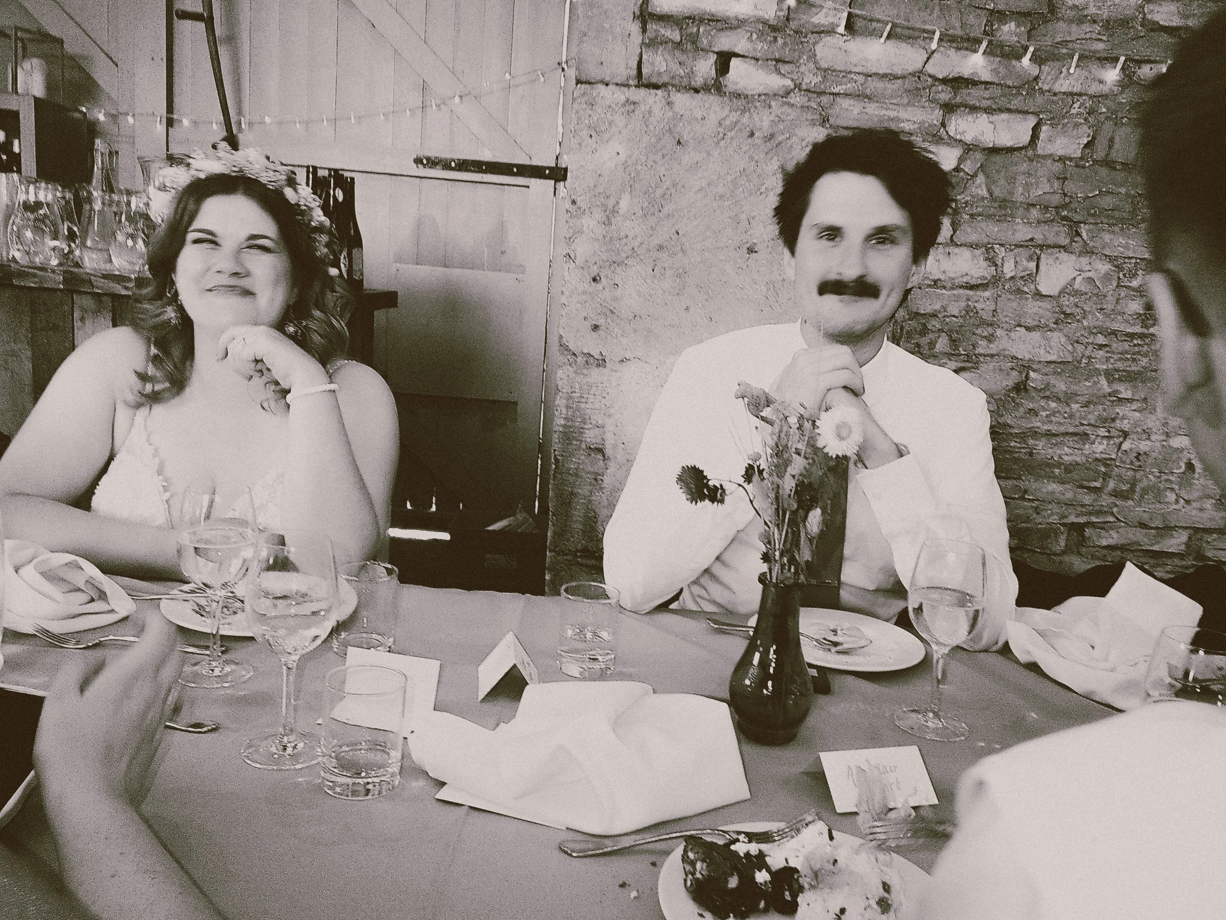 A black and white photo of a couple sitting at a dining table during a celebration. The woman on the left is smiling with a flower crown on her head, and the man on the right has a mustache, floral shirt, and is resting his chin on his hands, with a 