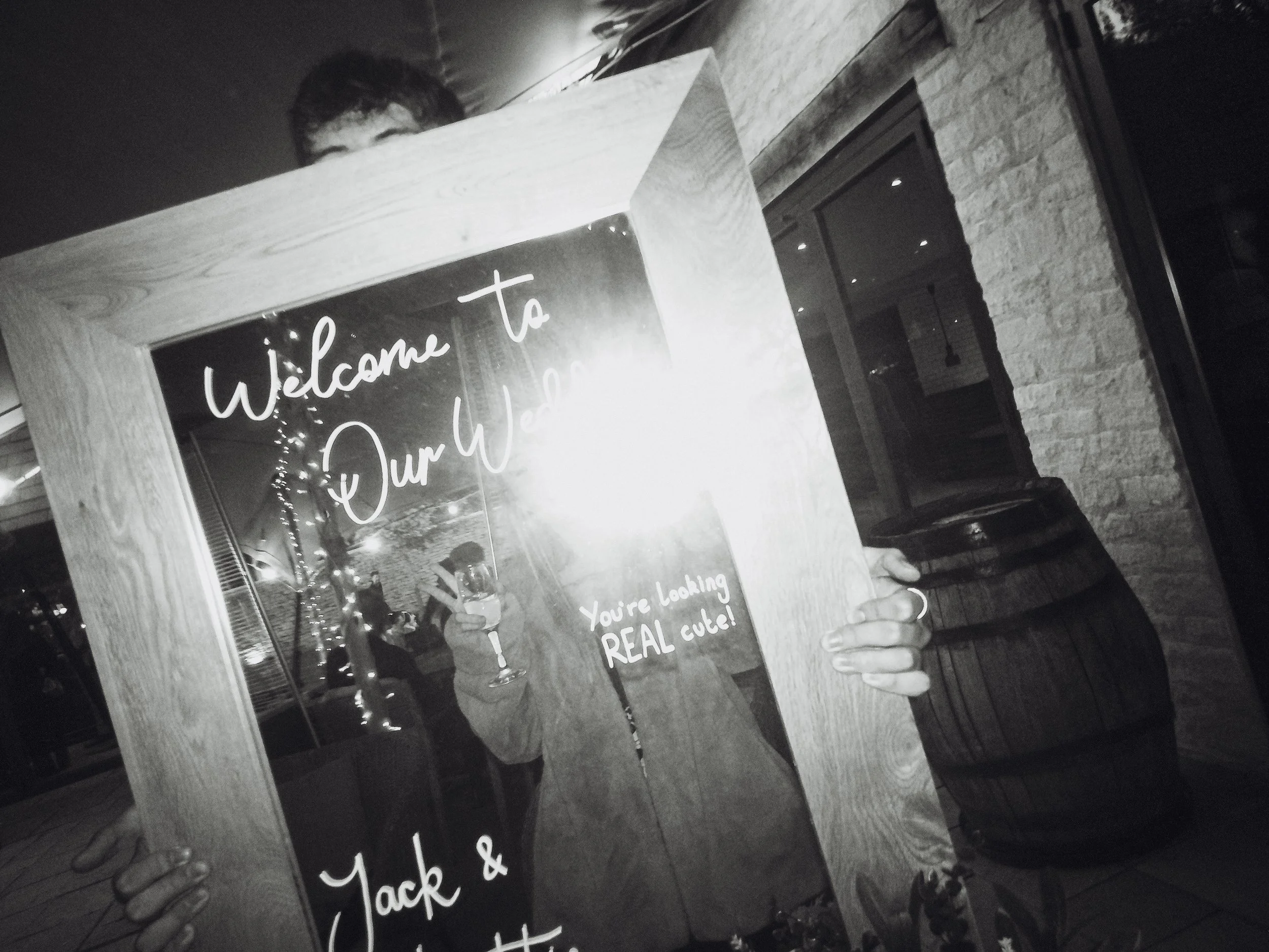 Person holding a mirror with a welcome message written on it, partially obscuring their face, in a cozy indoor setting with wood and brick decor, and people in the background