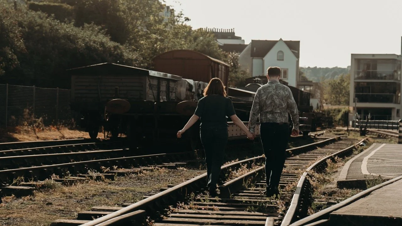 A man and woman walk hand-in-hand along railroad tracks in an urban area, with buildings and trees in the background.