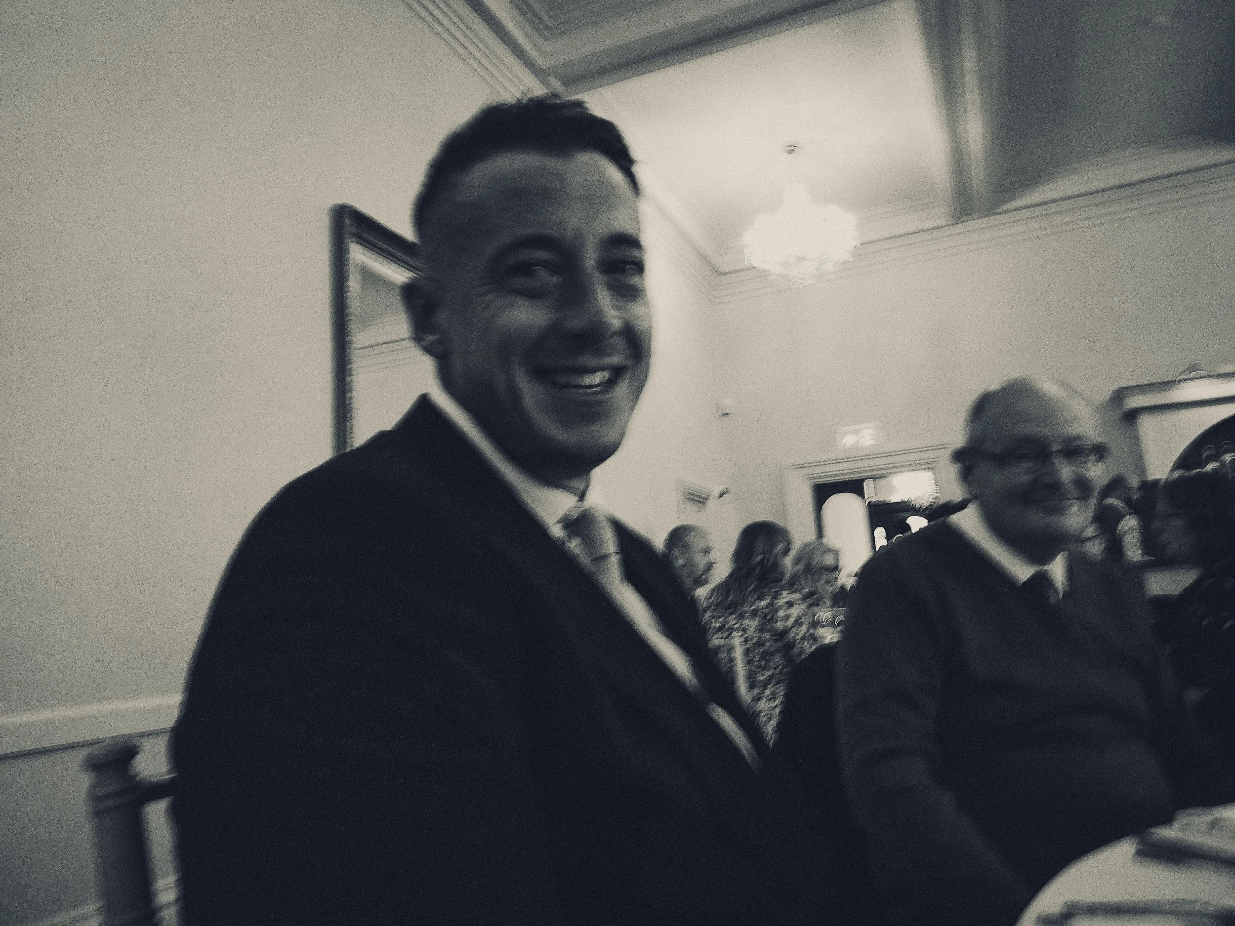 Two men sitting at a table, smiling, in a formal setting, with more people in the background.