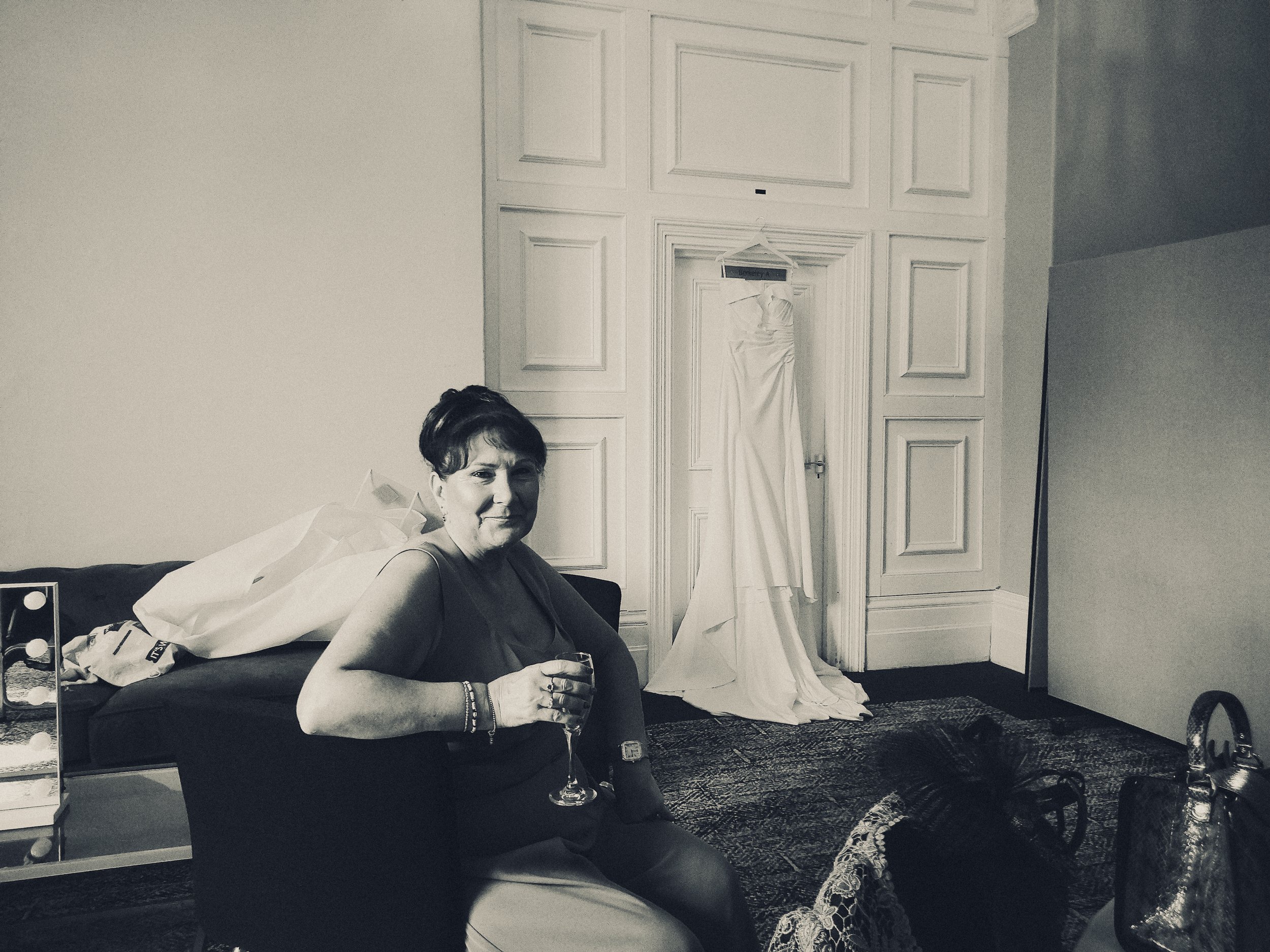 A woman sitting on a sofa holding a glass, with a wedding dress hanging on the door behind her in a room with patterned carpet and paneled walls.