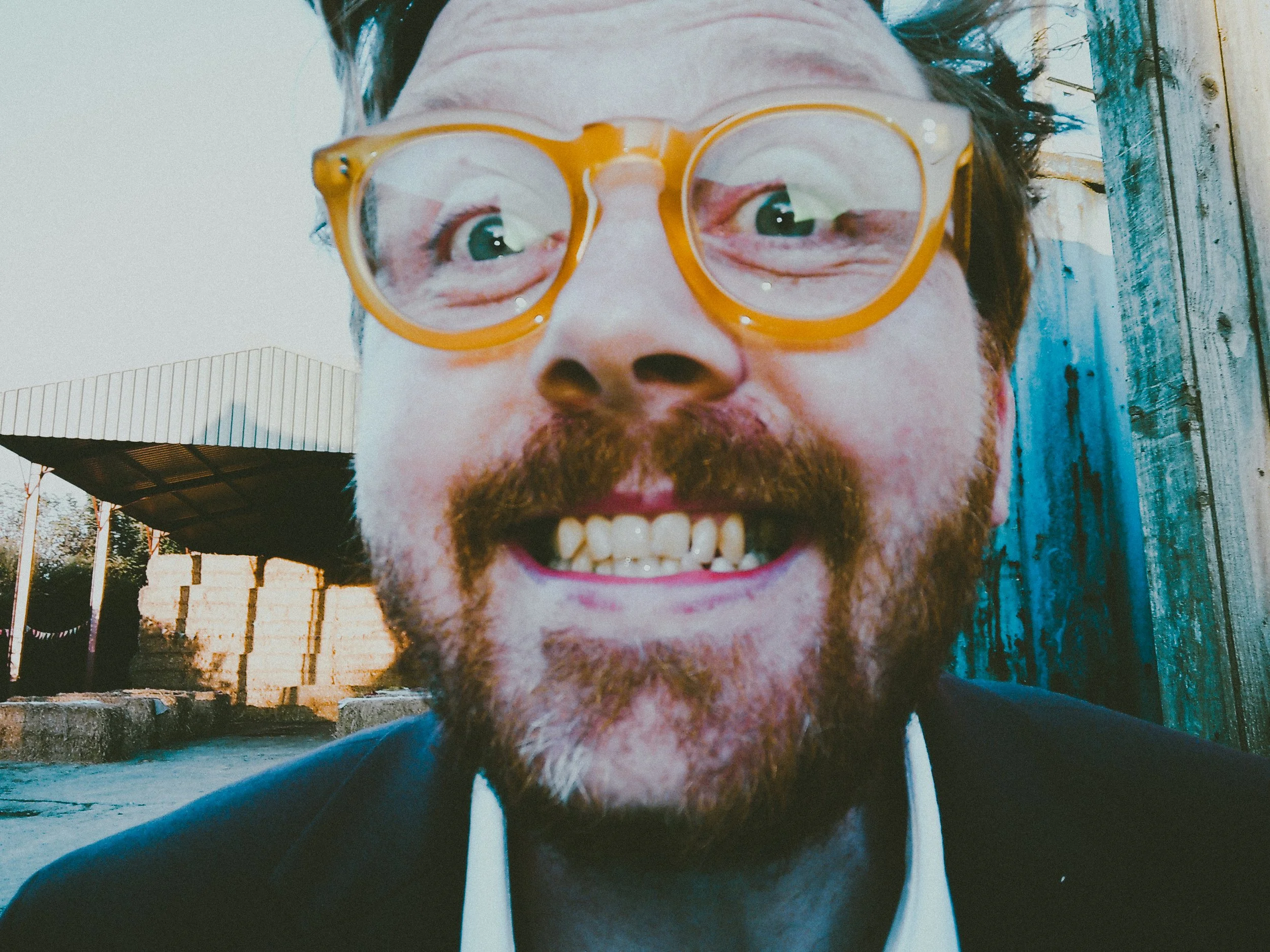Close-up of a smiling man with curly hair, glasses, and a beard, outdoors near a wooden fence.