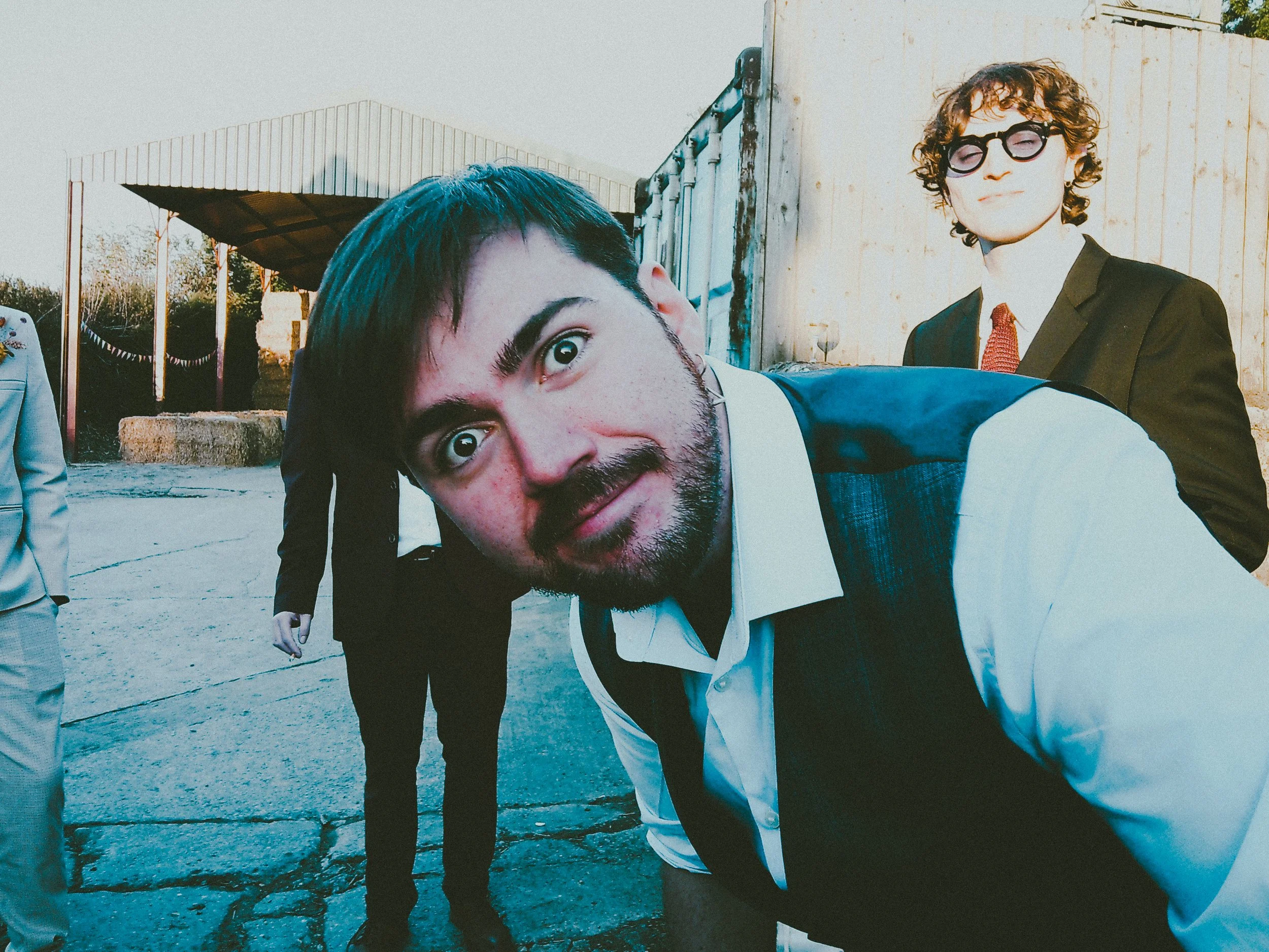 Close-up of a young man with curly hair and glasses in a suit. Another man with dark hair, a beard, and a vest leans toward the camera. People in suits are in the background outdoors.