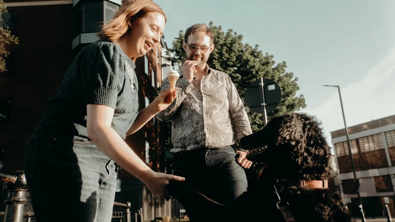 A young woman is holding an ice cream cone and smiling, while a man in glasses and a patterned shirt looks at her and a large black dog is sitting in front of her on a leash. They are outdoors on a sunny day with buildings and trees in the background