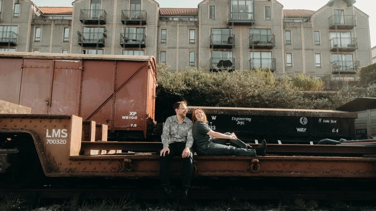 Two smiling people sit on a flat train car, with a building and plants in the background.