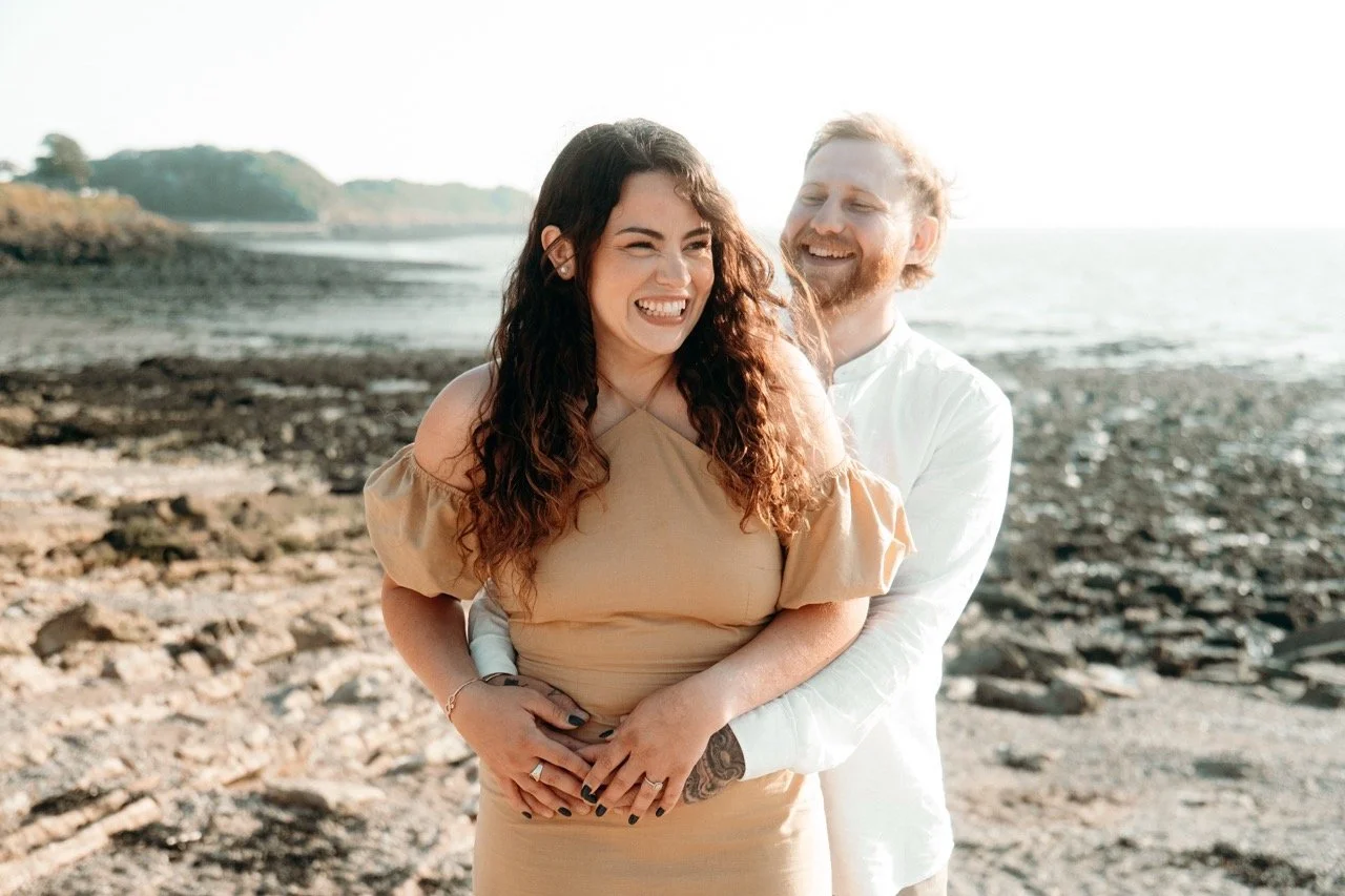 A happy couple on a rocky beach, smiling and embracing each other. The woman has long, curly brown hair and is wearing a beige dress with off-shoulder sleeves. The man has short, light-colored hair and a beard, wearing a white shirt. They are enjoyin