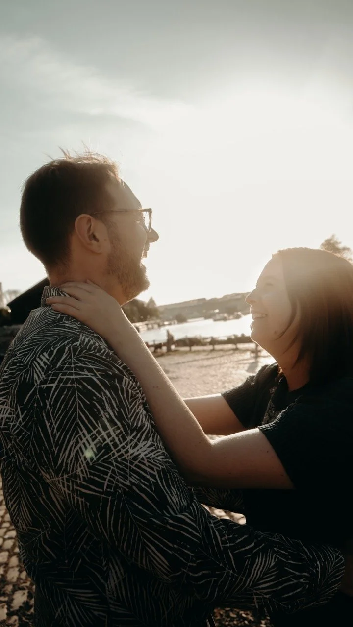 A couple smiling and embracing outdoors near a body of water at sunset, with warm sunlight illuminating their faces.