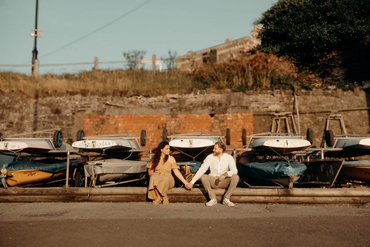 A couple sitting on the ground holding hands near a row of boats turned upside down, with a brick wall and trees in the background.