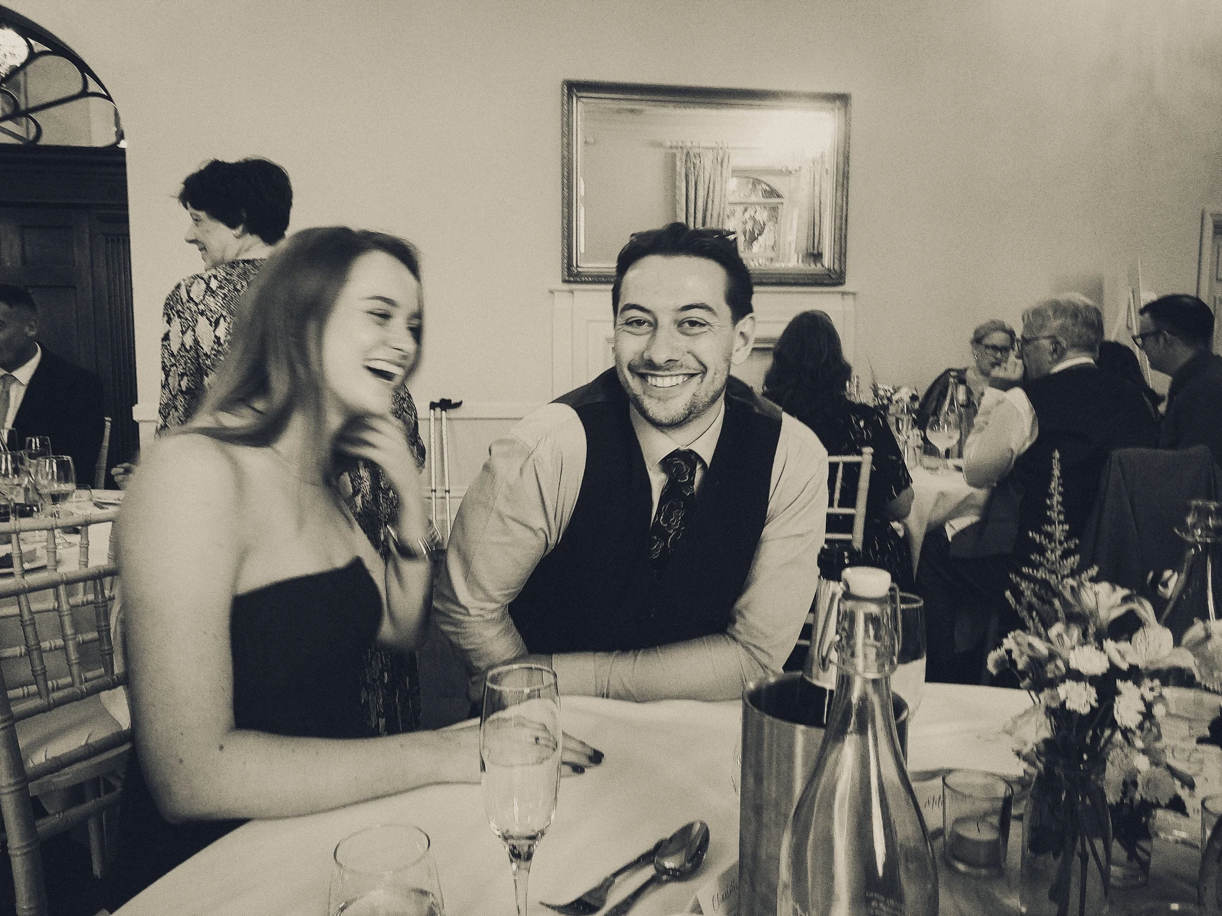 People smiling and chatting at a formal event or dinner in a decorated room with a mirror, table settings, and flowers.