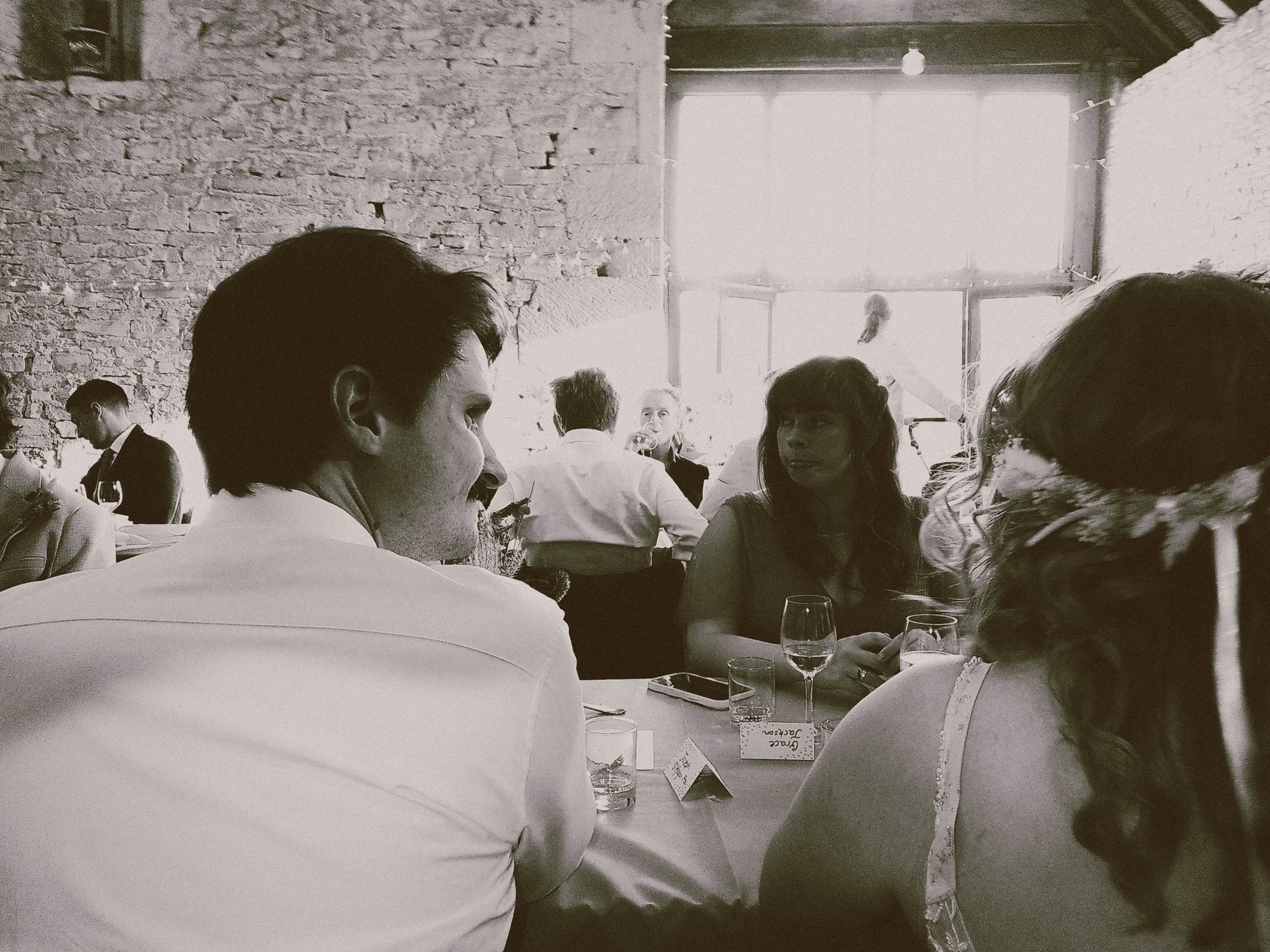 People sitting at a table in a rustic-style venue with brick walls and large windows, engaged in conversation.