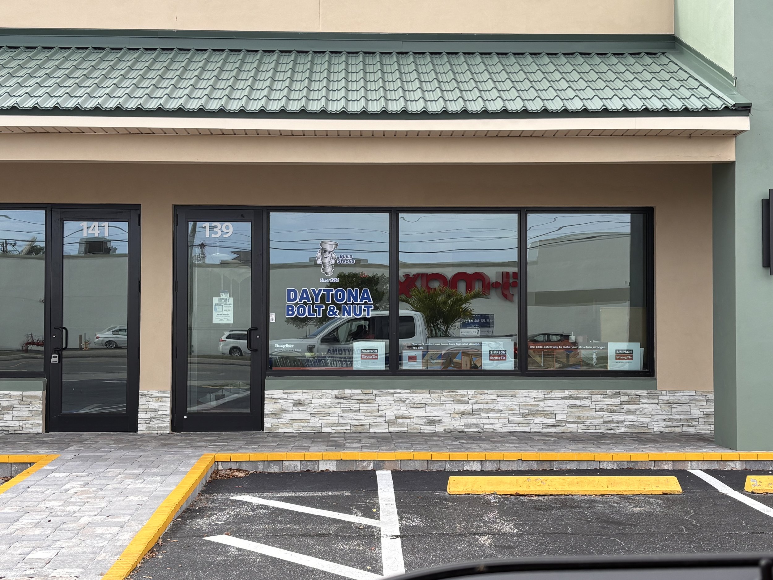 Exterior view of a storefront with large glass windows and two glass doors, with a parking lot in front. The store sign reads 'DAYTONA BOLT & NUT' and includes a logo with a hammer character. There are parked vehicles and a building with a green roof can be seen reflected in the windows.