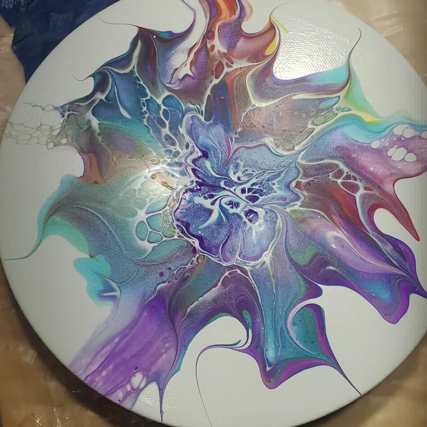 Abstract acrylic pour painting with swirling colors including purple, blue, green, red, and white on a round canvas.