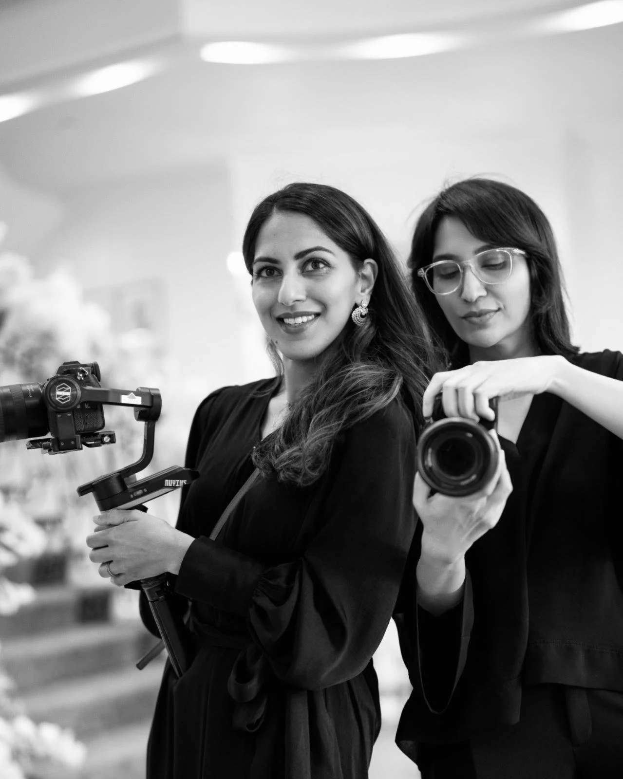 Two women with cameras taking a selfie indoors, smiling and one with her eyes closed.