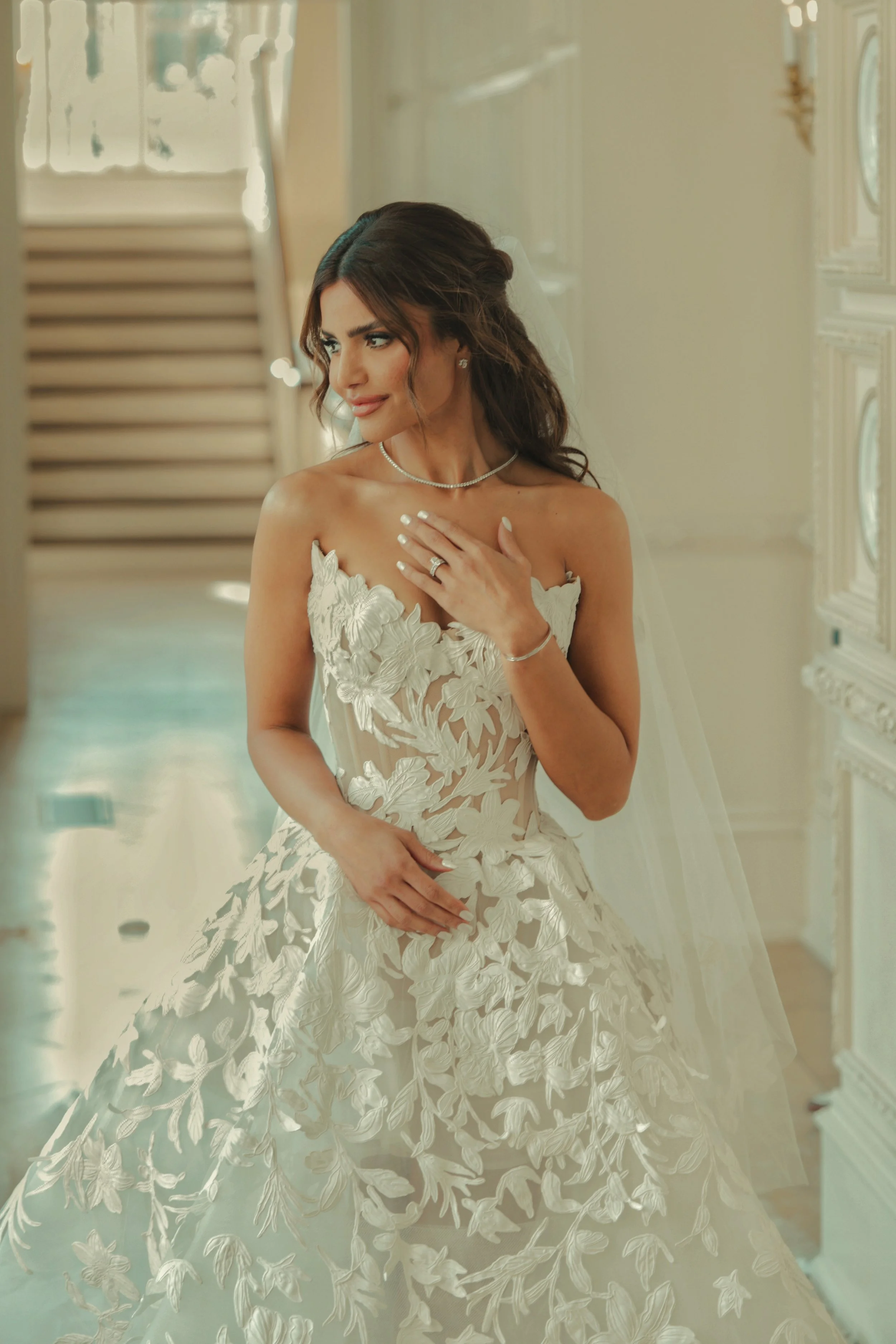 A bride in a white wedding dress with floral lace embroidery, standing in an elegant room with beige walls and wooden shutters, with a gentle expression and her hand on her chest.