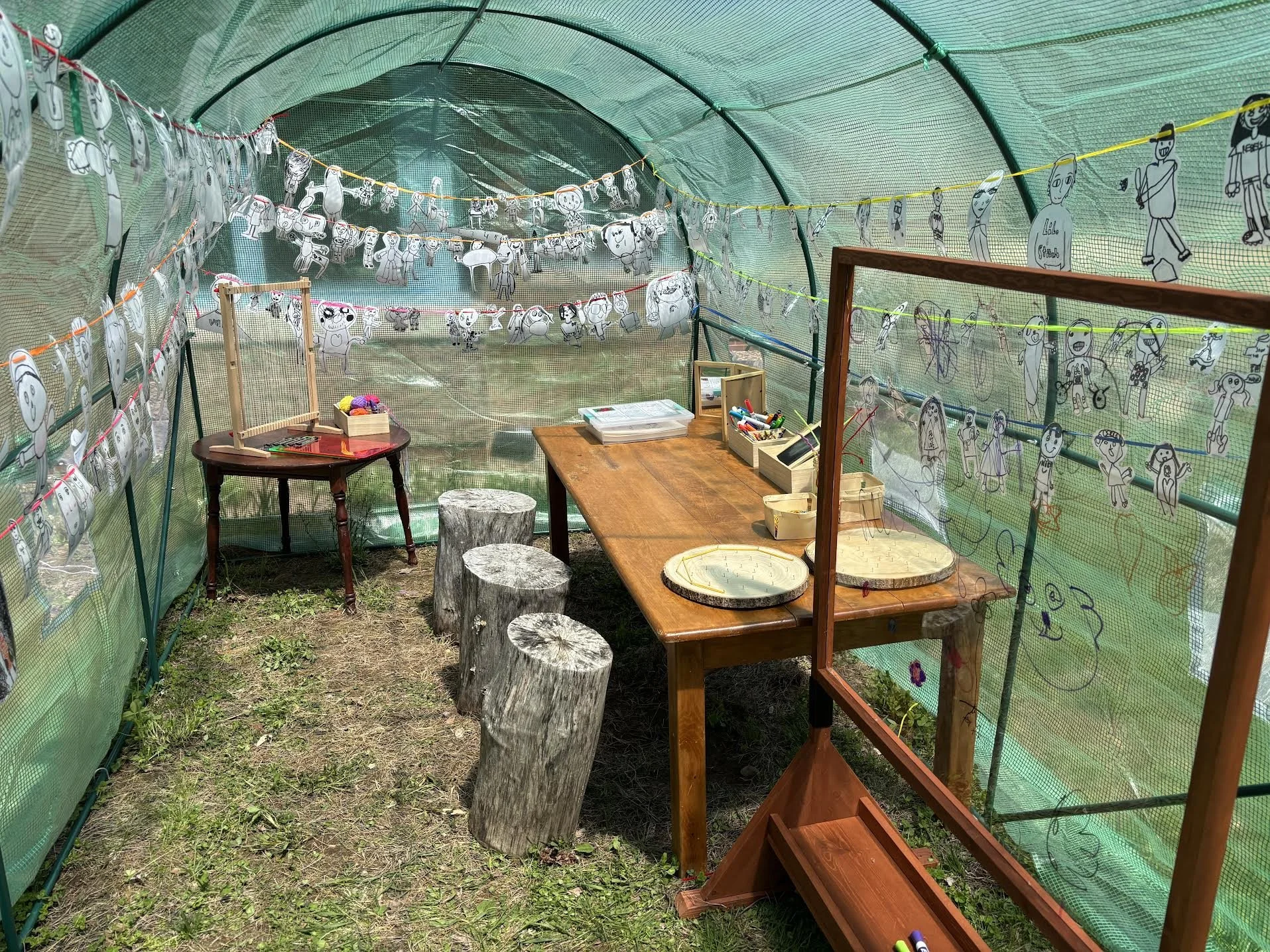 A greenhouse with a table, three stools, and a clear chalkboard inside with elementary school artwork hanging on string inside on the walls on a sunny day