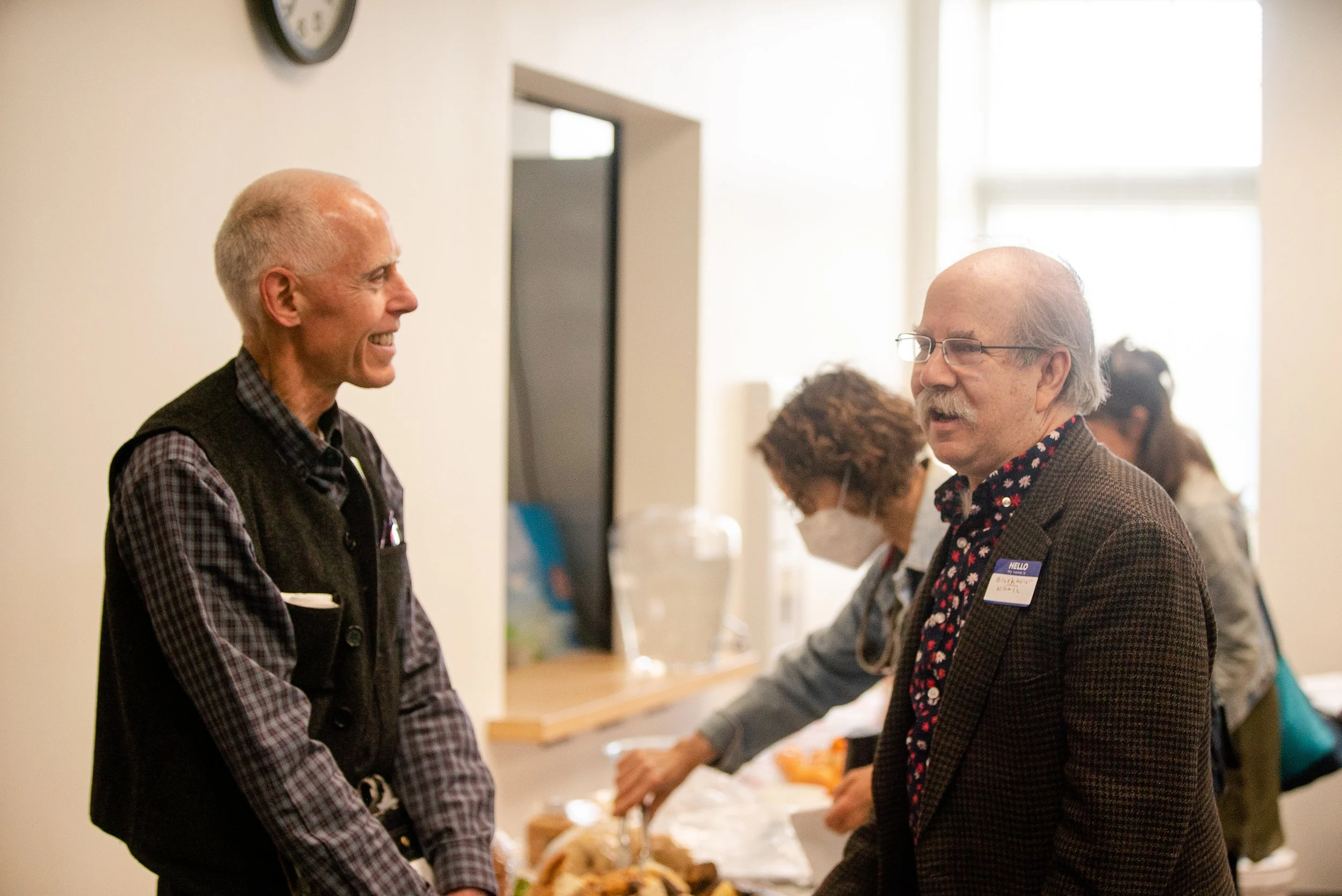 Two older adult men smiling and talking while facing each other inside of a conference room