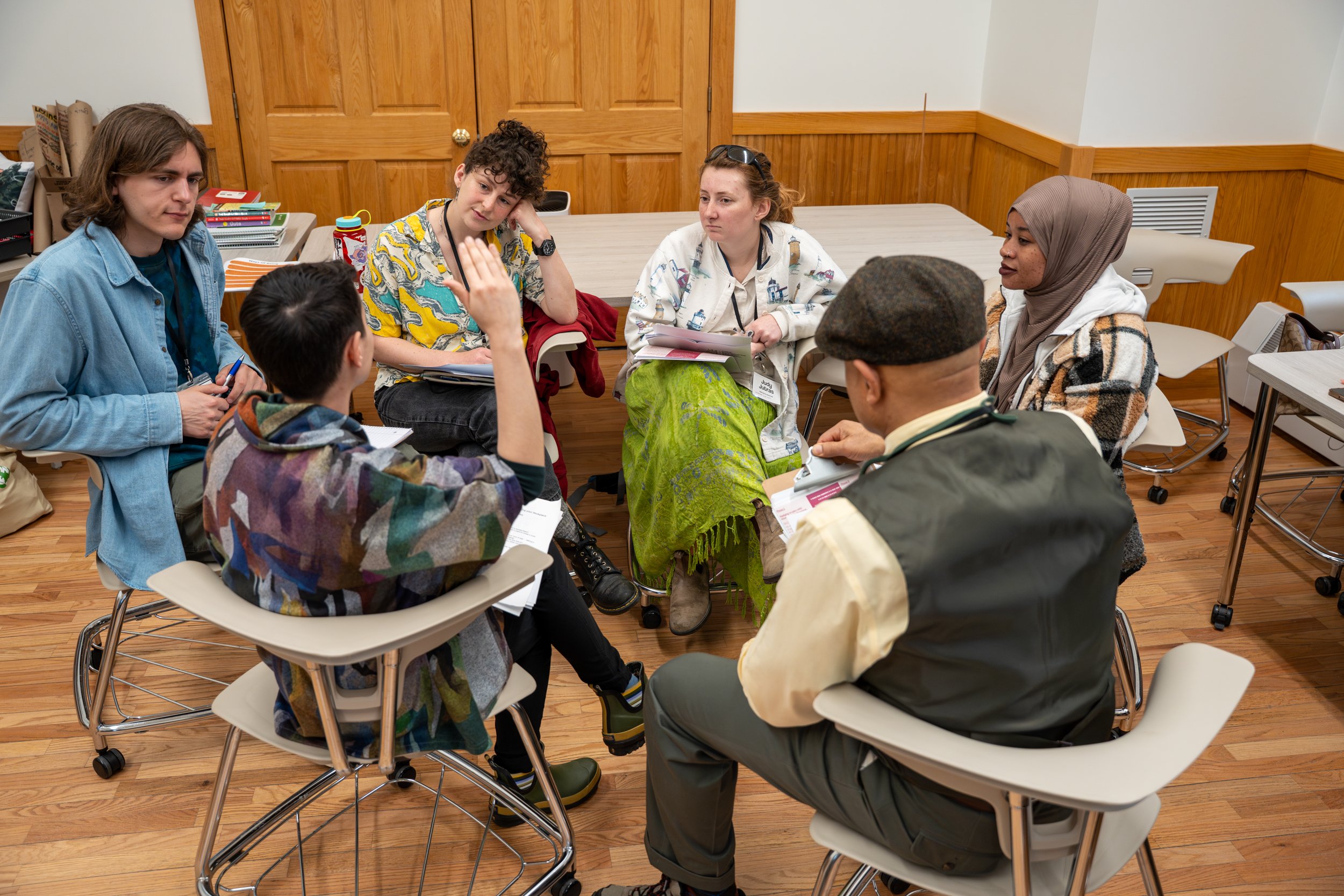 Six people sit in school desks formed in a circle inside of a classroom discussing.